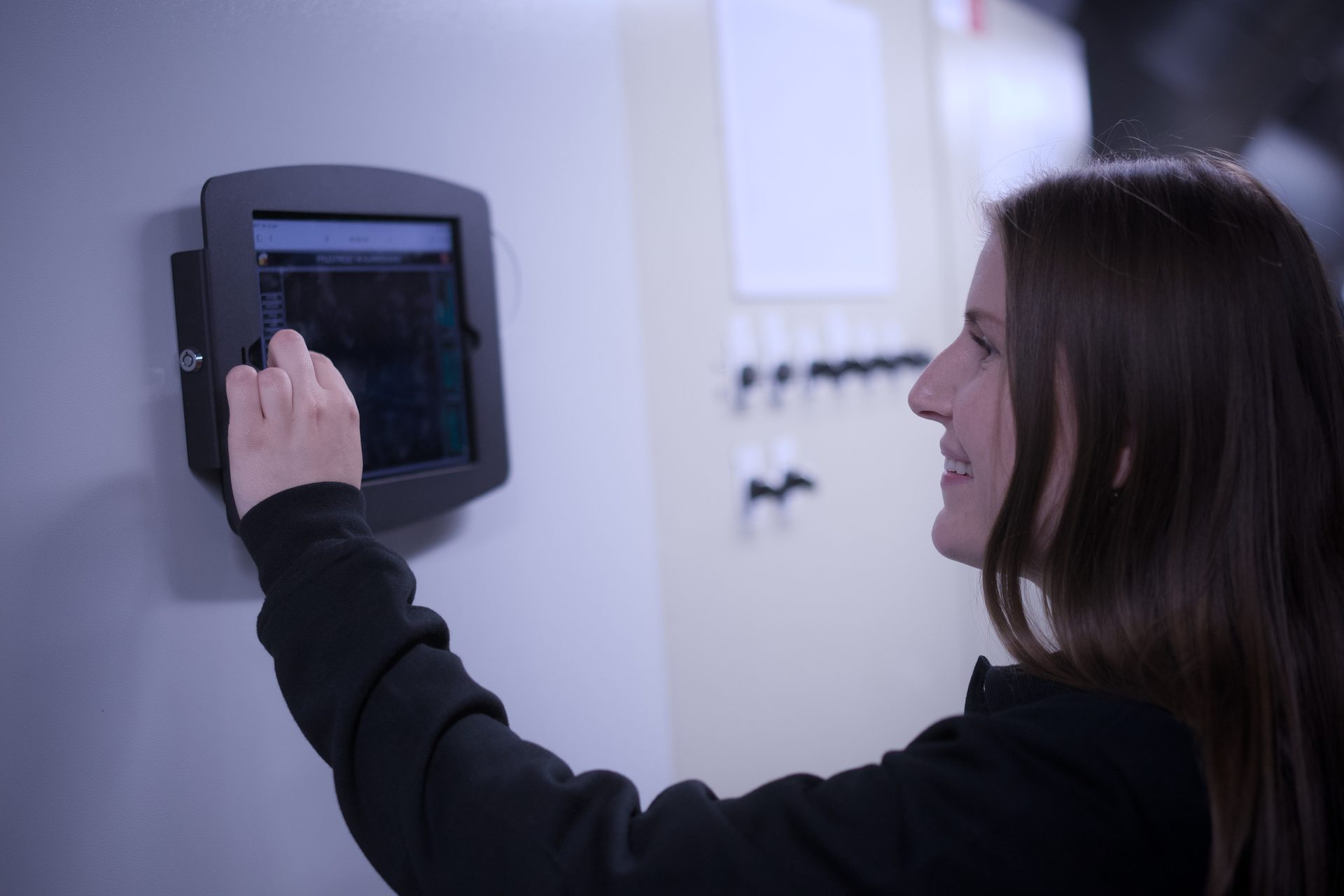 Woman interacts with a wall-mounted tablet, possibly for industrial or technical control, smiling in profile.