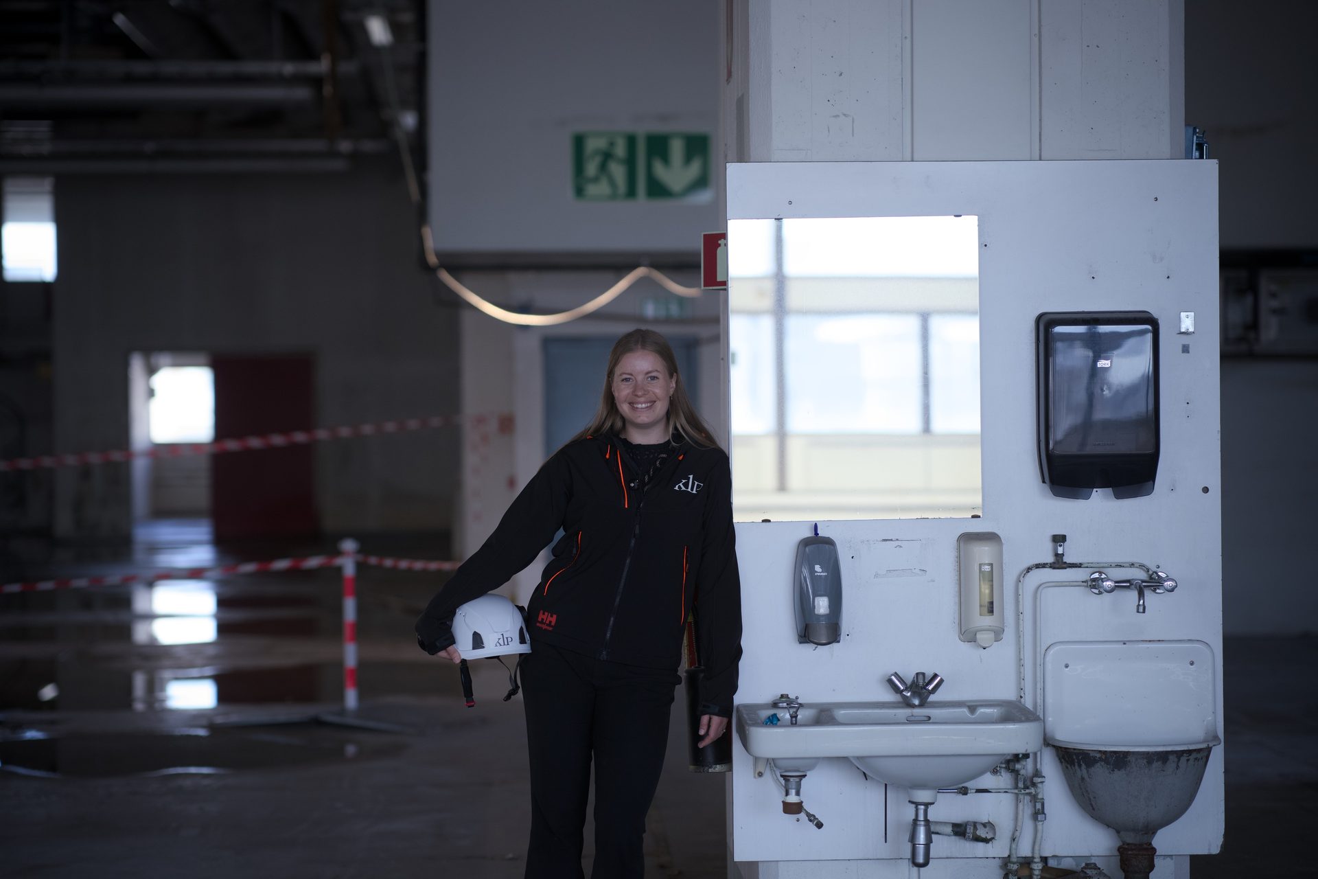 Smiling woman in work jacket holding a hard hat next to a temporary sink station in an industrial building.