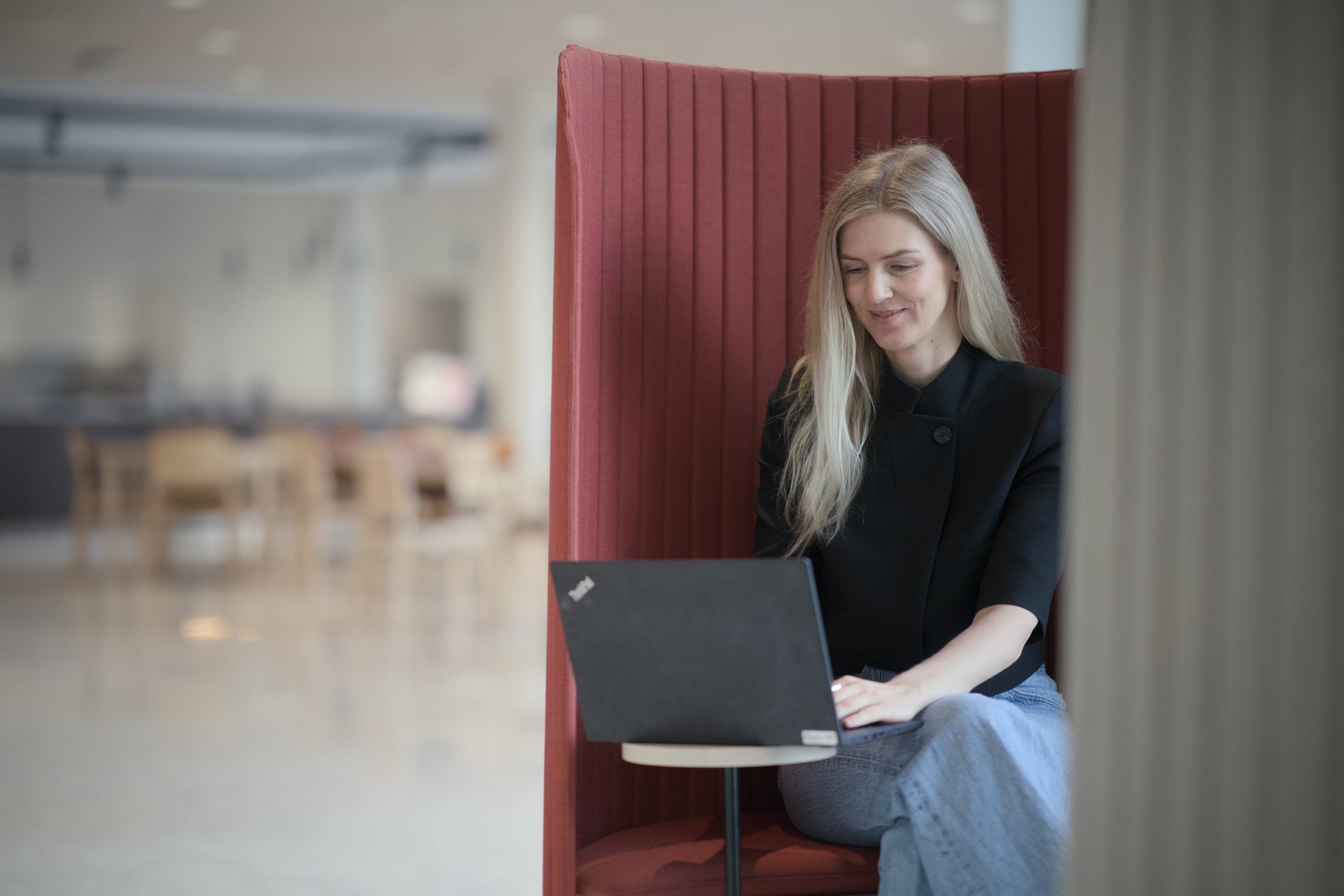 Woman with blonde hair working on a laptop in a red booth chair in a modern office.