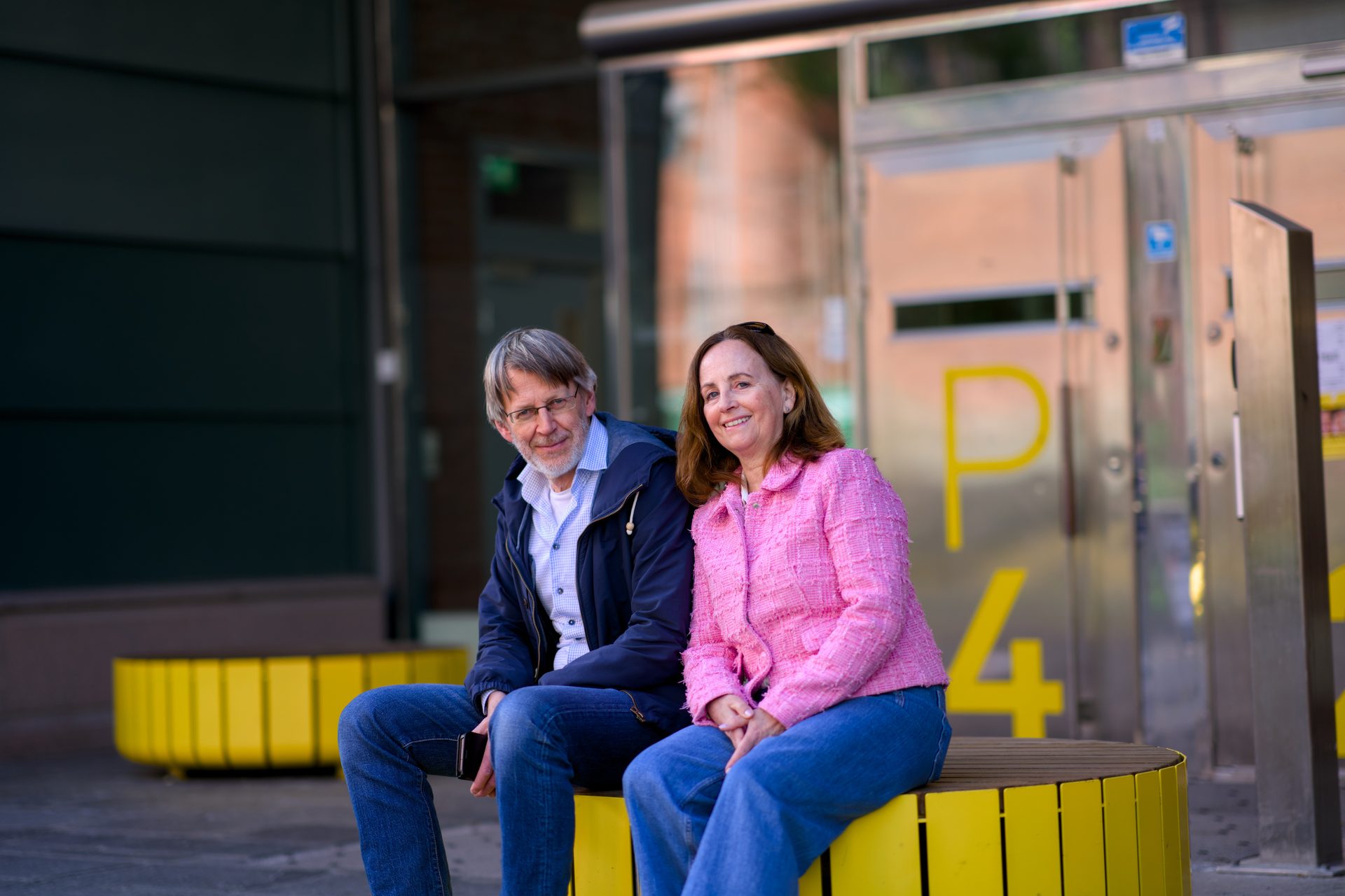 Two smiling people, a man and a woman, sitting on yellow benches in an urban setting.