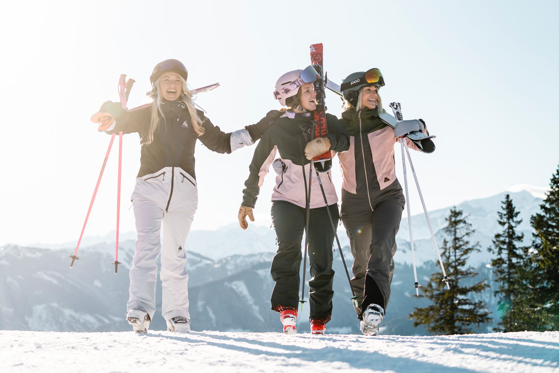 Three smiling women carrying skis and poles on a bright, snowy mountain slope.