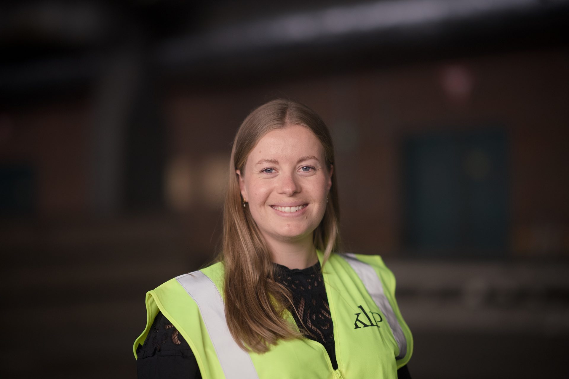 A smiling young woman in a yellow high-visibility vest with long blonde hair looking at the camera.