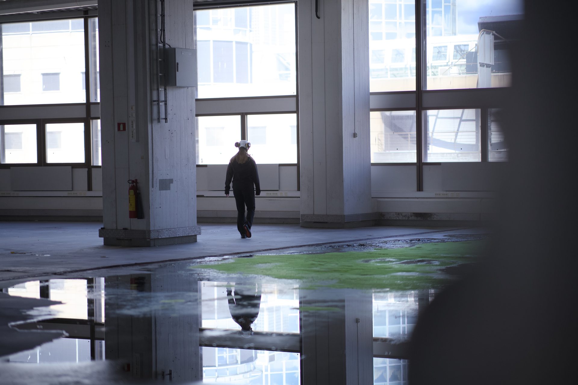 A person in a respirator walks through an empty, reflective industrial space with large windows and puddles.