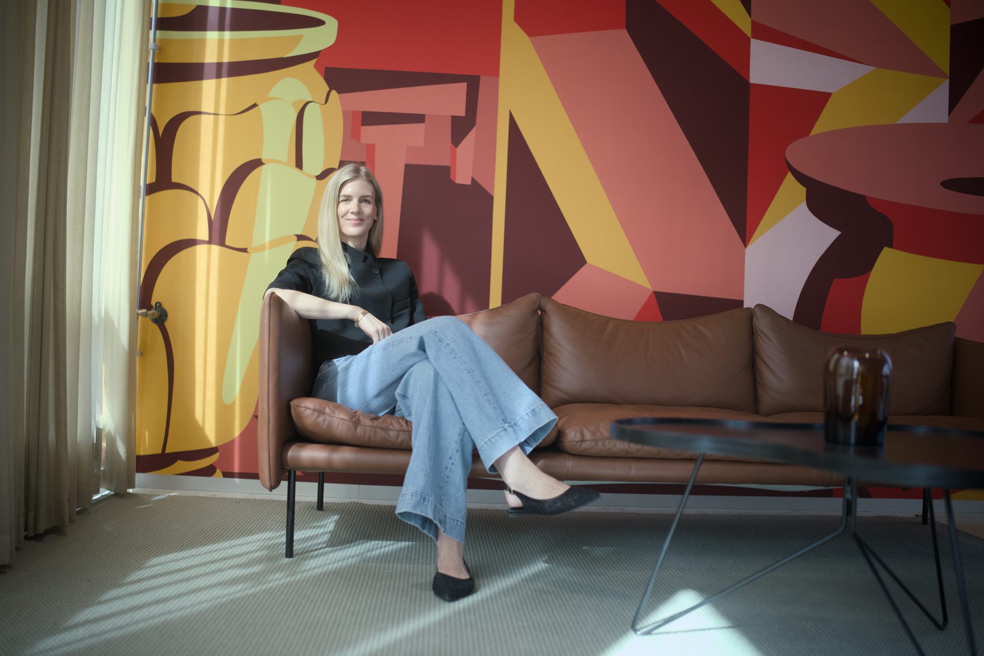 A woman with blonde hair in a black shirt and jeans sits on a brown sofa in a room with a colorful mural.