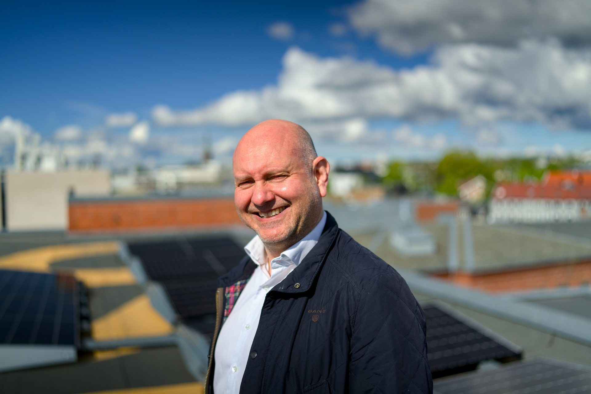 Bald man smiling on a rooftop with solar panels and city in the background under a blue sky.