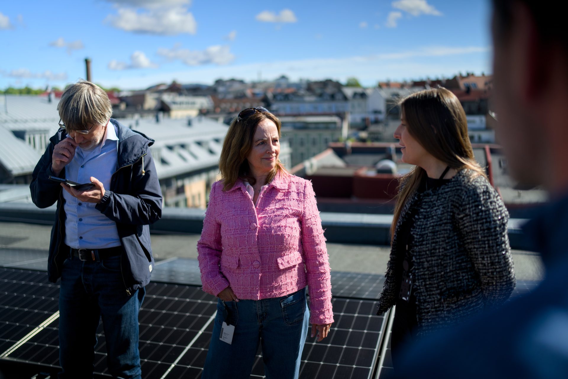Three people on a rooftop with solar panels, two women conversing, and a man looking at his phone.