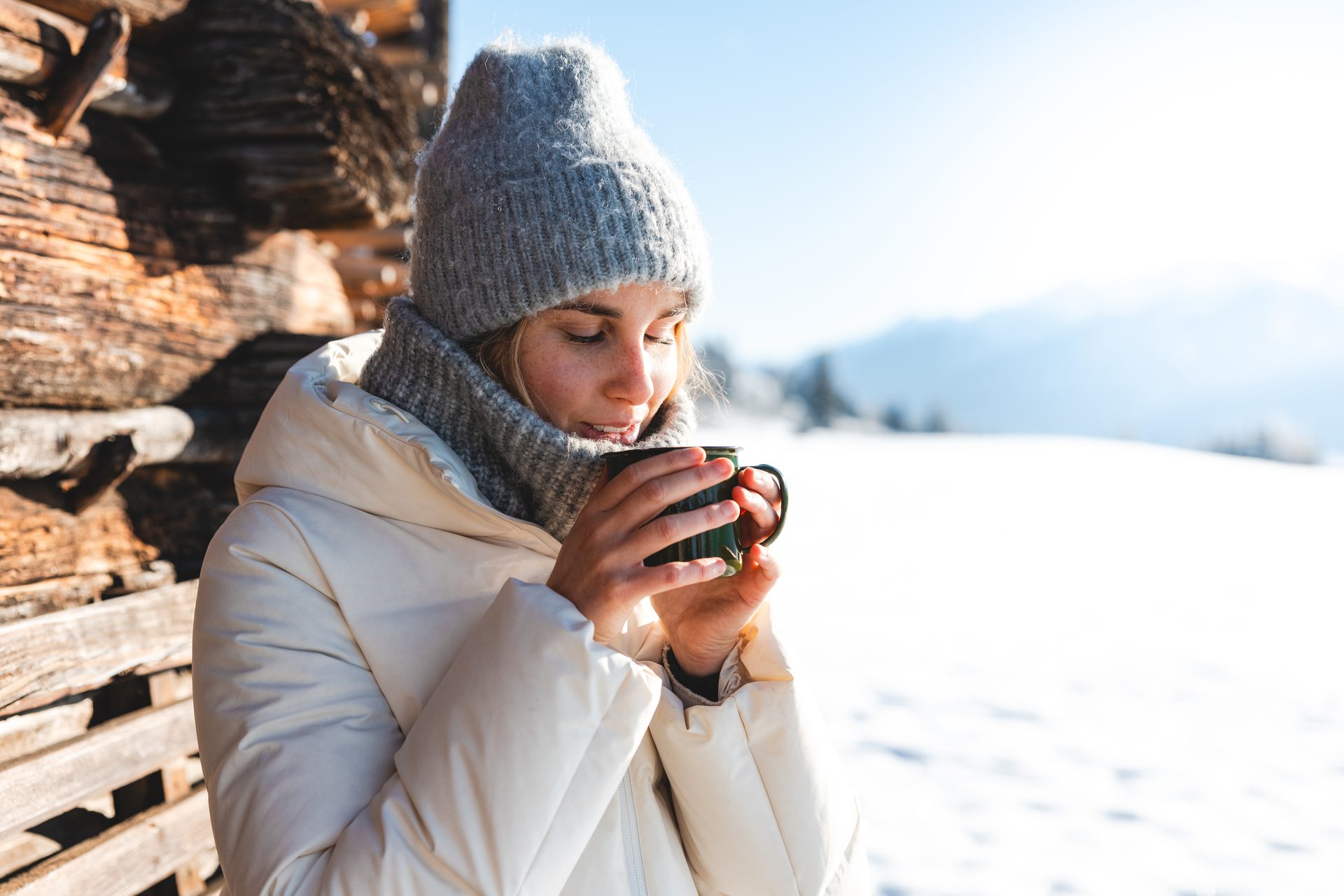 Woman in winter clothes enjoying a hot drink from a green mug in a snowy mountain landscape.
