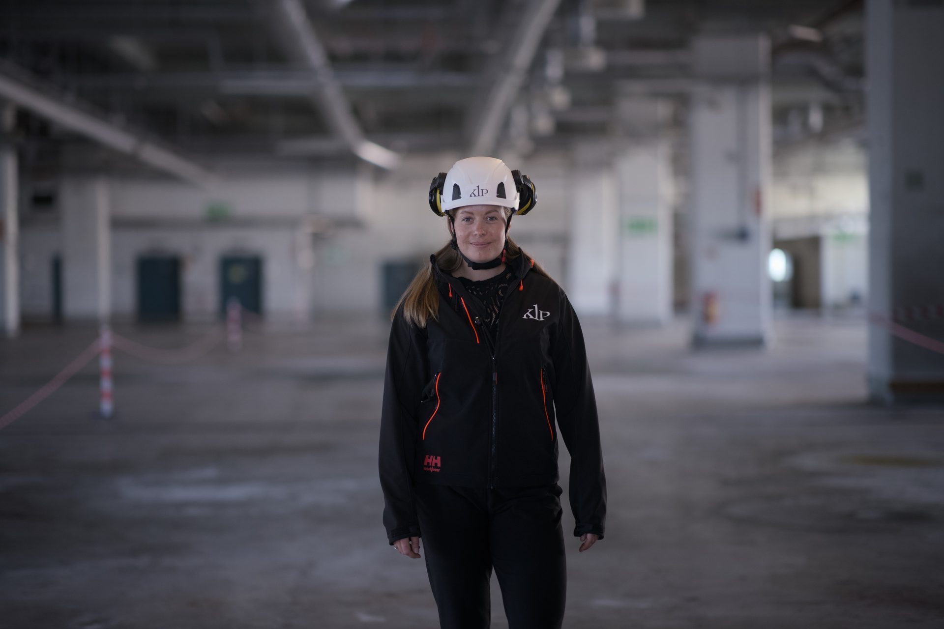 Woman in hard hat and work jacket smiling in a large, empty industrial space.