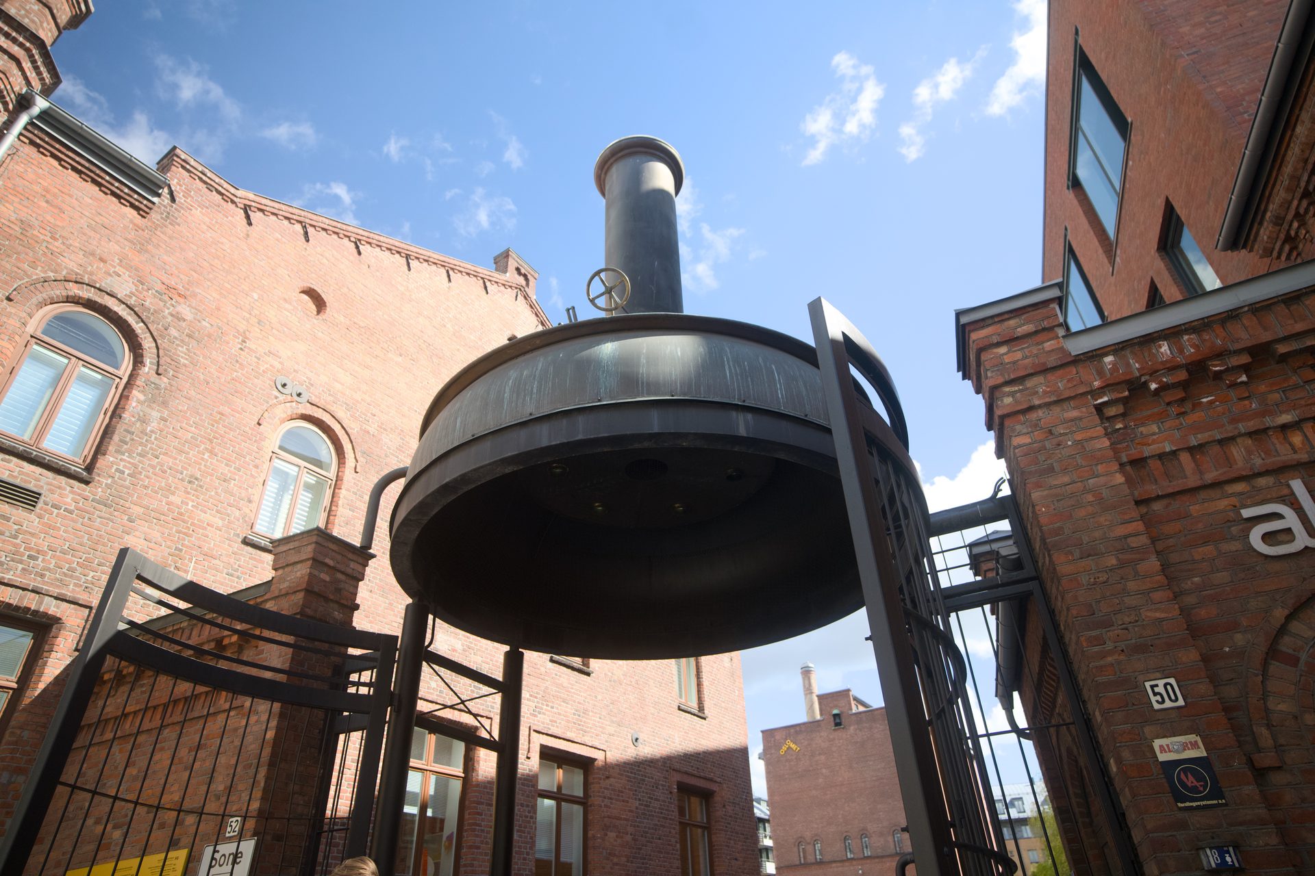 A low-angle view of a large, dark industrial vat with a stack, surrounded by old red brick buildings.