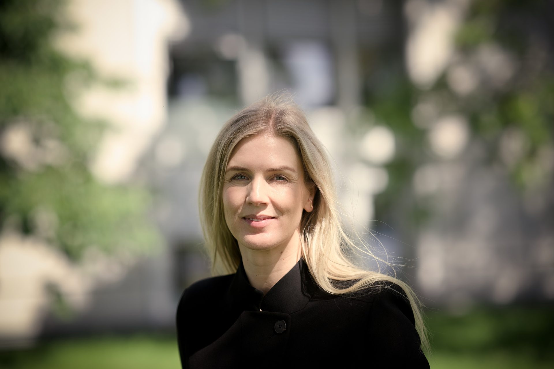 A smiling blonde woman in a black jacket, outdoors with a soft focus background.