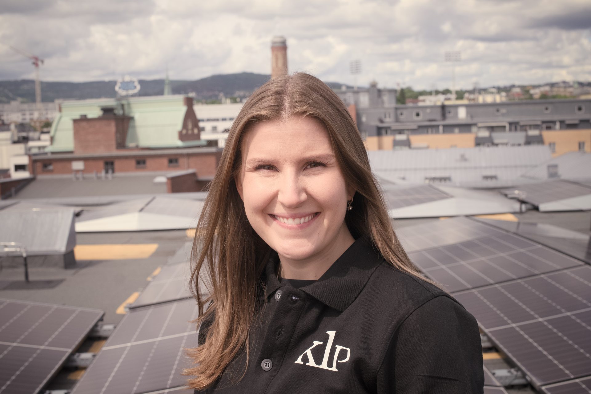 A smiling woman in a black KLP shirt stands on a rooftop with solar panels and a city skyline.