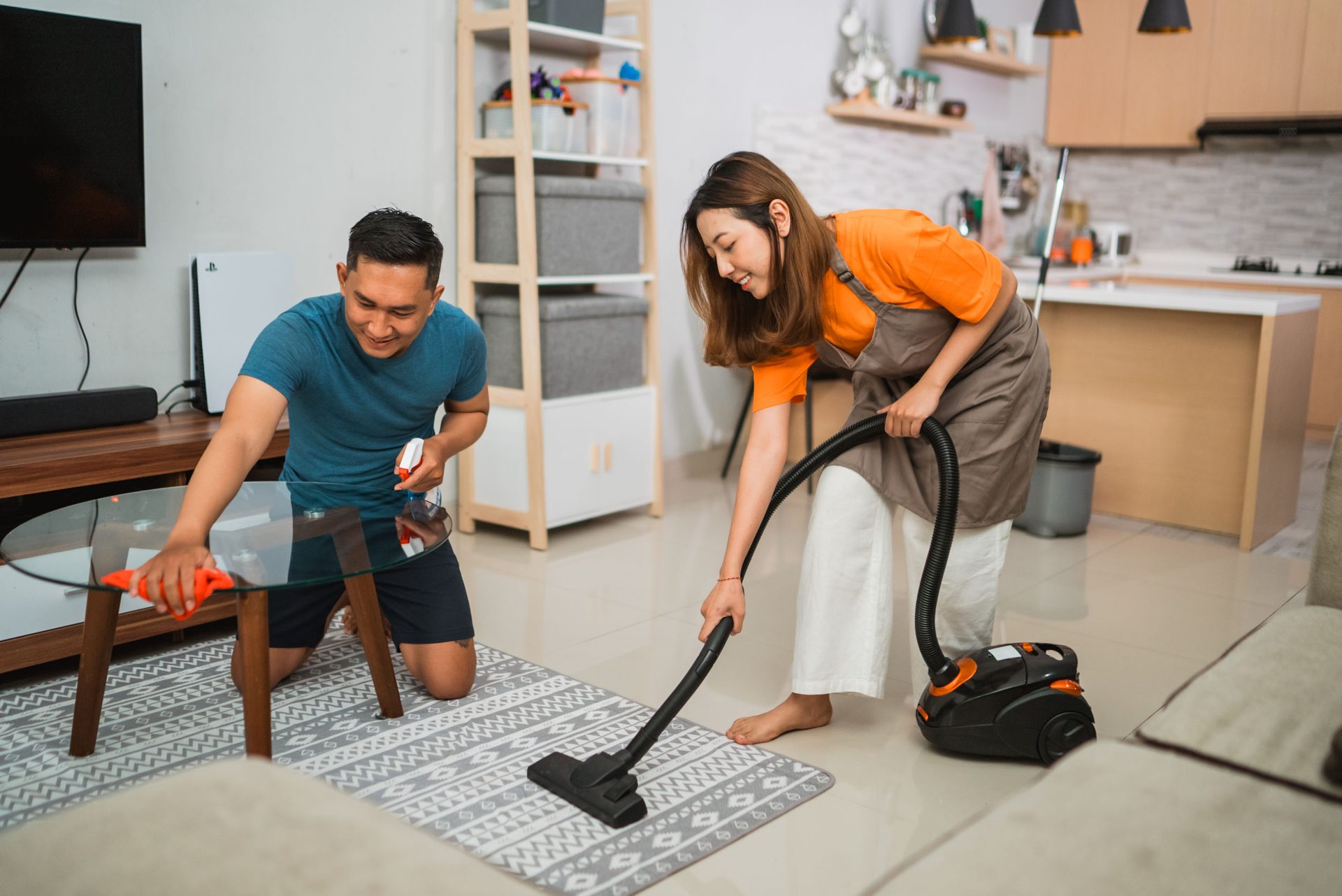 Couple cleaning living room: man wiping table, woman vacuuming.