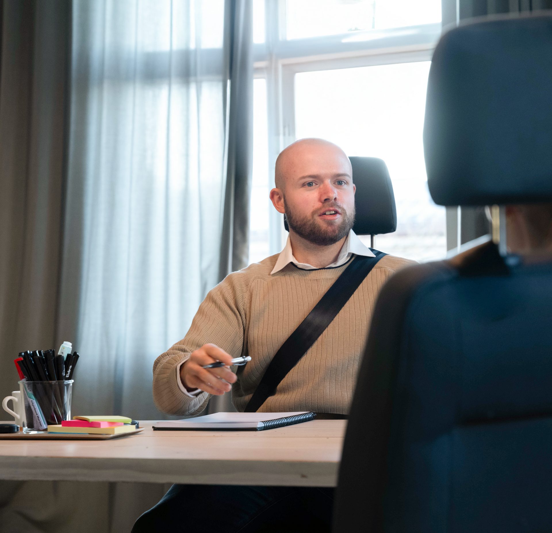 A bald man with a beard, wearing a seatbelt, talks at a desk while holding a pen.