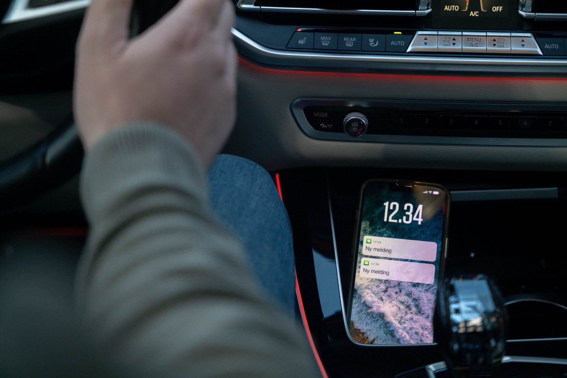Driver's hand on steering wheel, smartphone showing "Ny melding" notifications on a car's charging pad.
