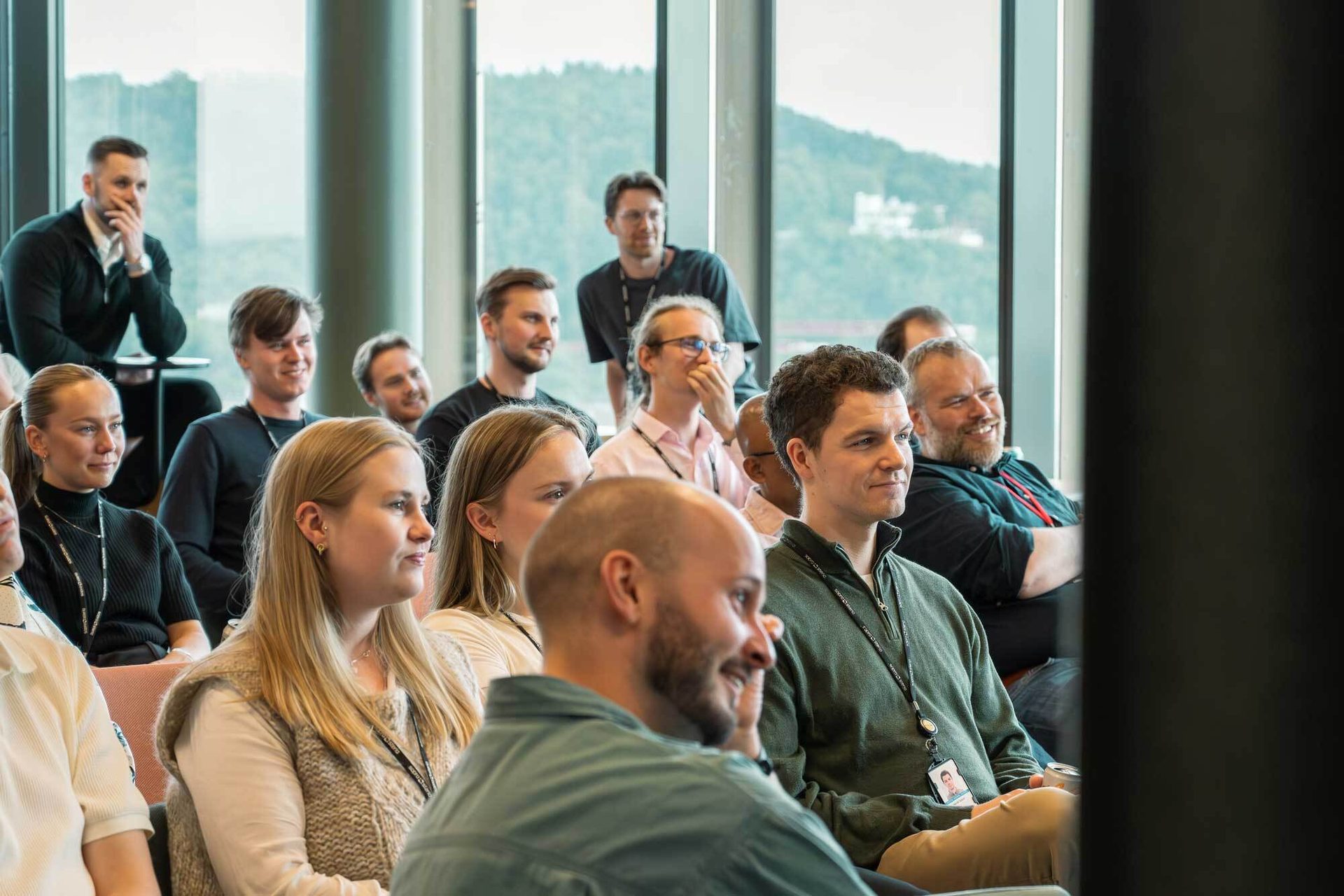 A group of diverse people in a modern setting, sitting, listening, and smiling during a presentation.