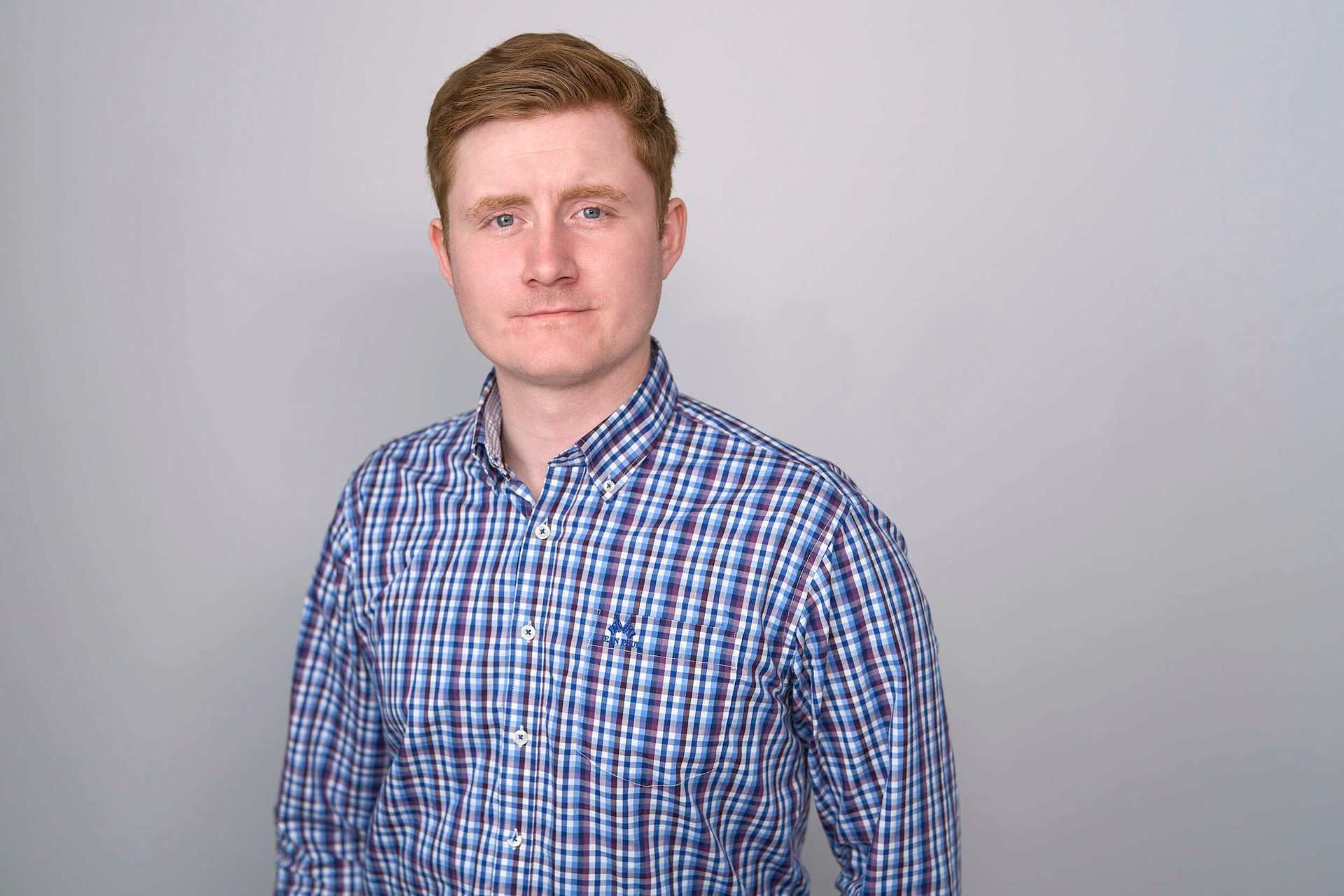 A man with red hair in a plaid shirt looks directly at the viewer against a gray background.