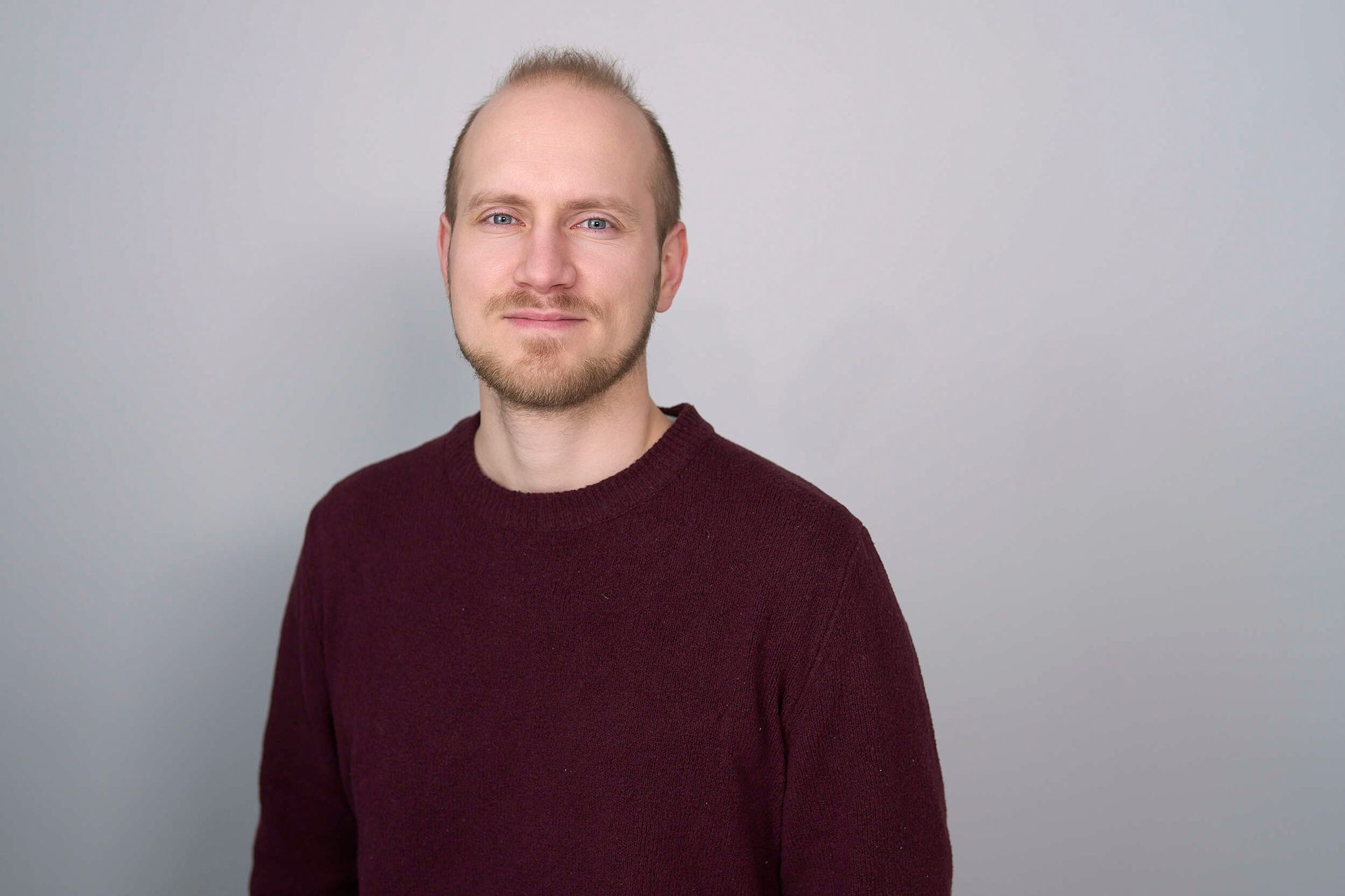 Portrait of a man with light hair and beard, wearing a maroon sweater, looking at the camera.