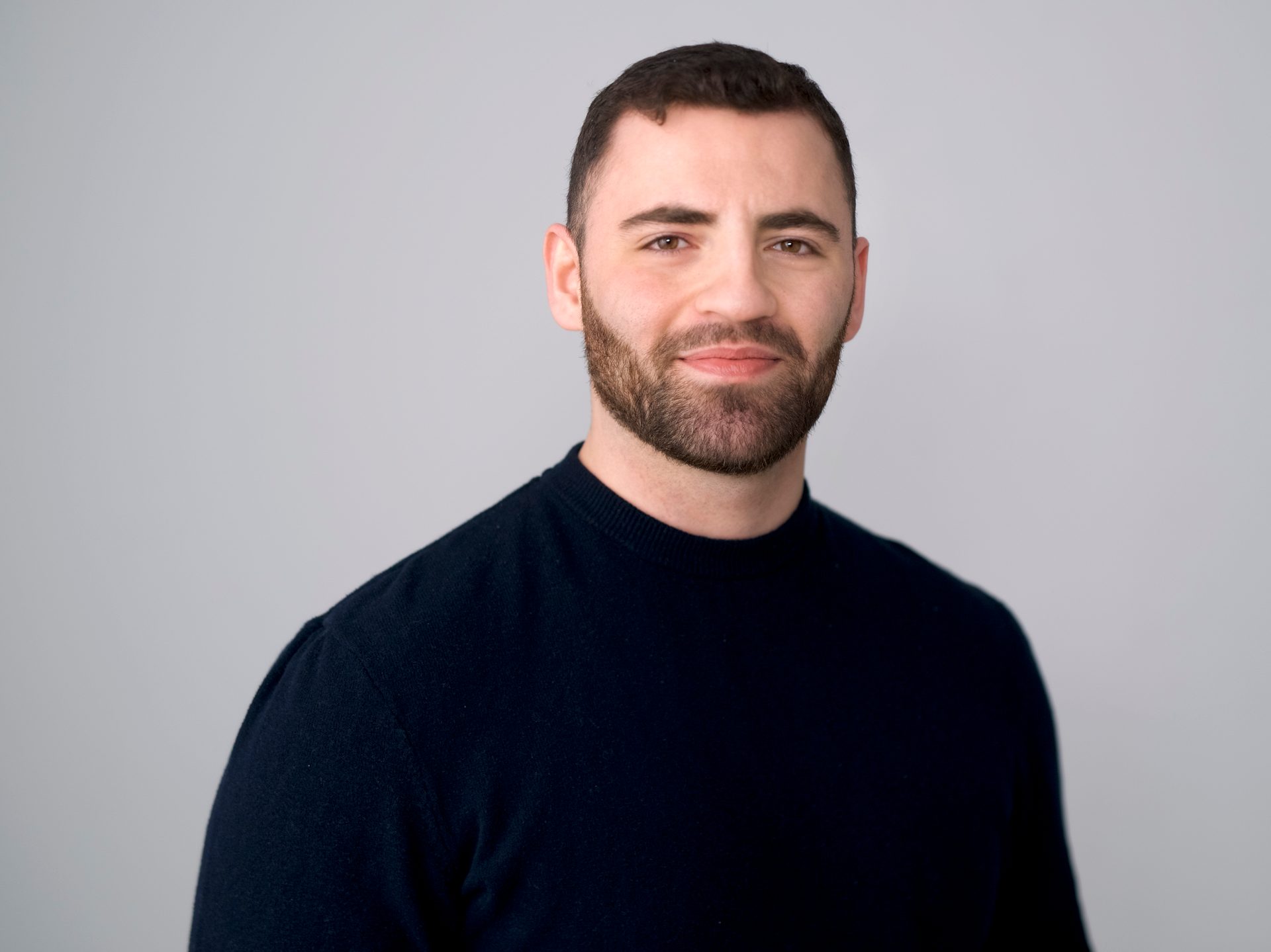 Headshot of a man with dark hair and a beard, wearing a dark sweater, against a light gray background.