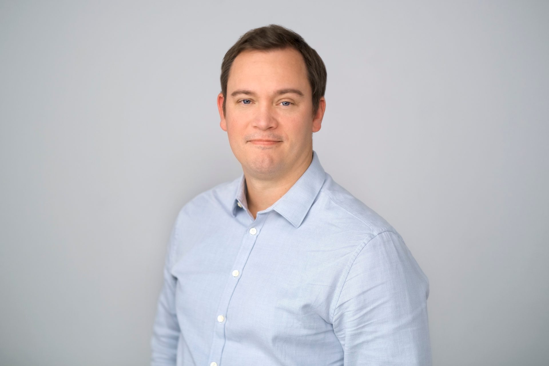 A man with short brown hair and blue eyes, wearing a light blue button-up shirt, smiles slightly.