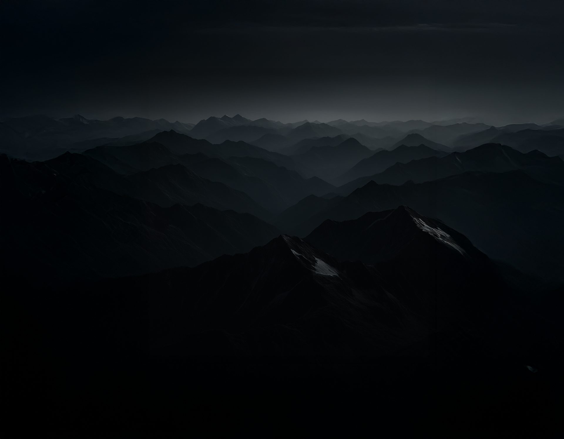 Dark, layered mountain peaks stretching into the distance under a gloomy sky, subtle light on some.