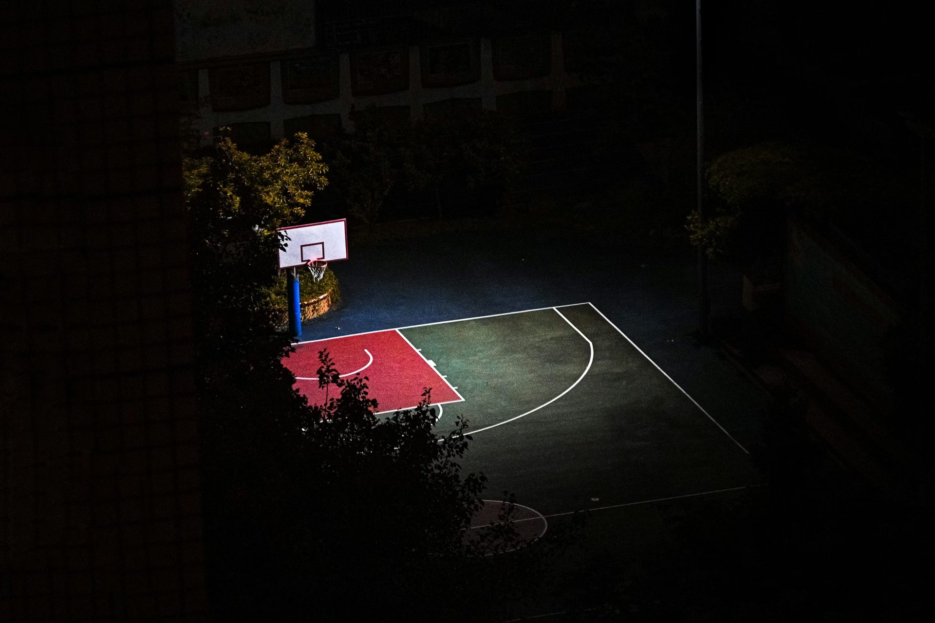 A partially lit basketball court at night, with a red key area and dark surface, viewed from above.