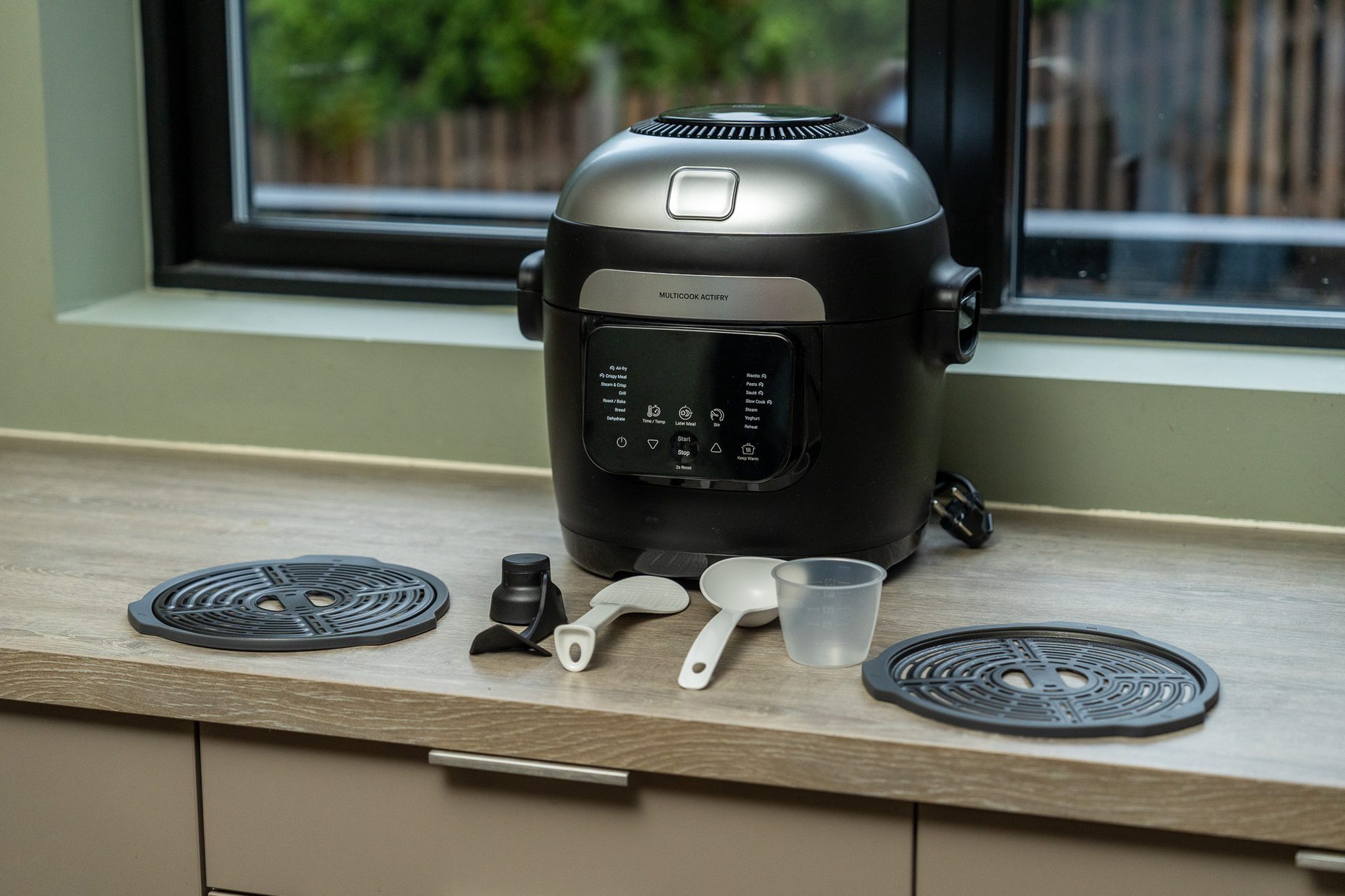Multicook Actifry appliance, black and silver, with cooking accessories on a kitchen counter.