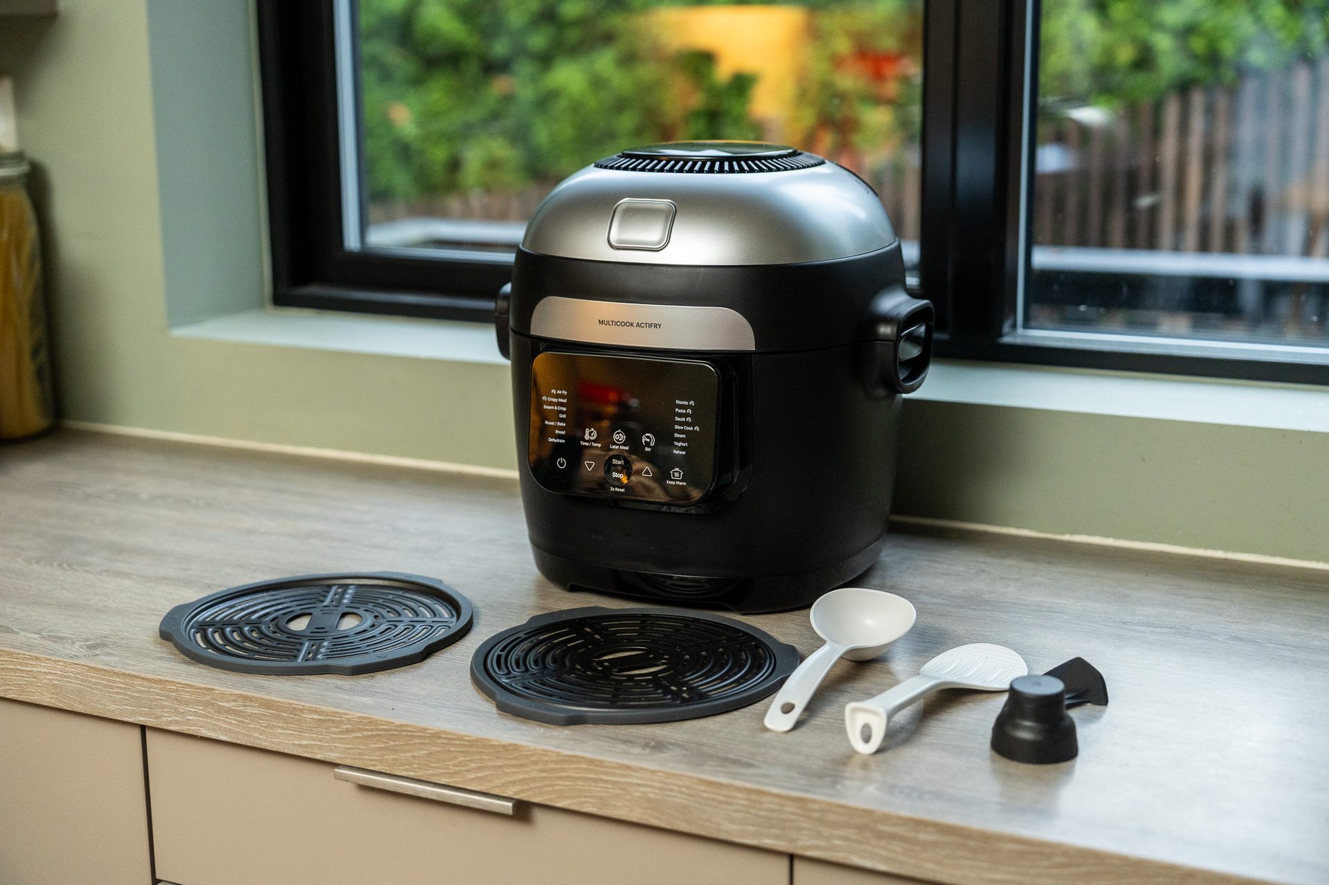 Black & silver multi-cooker with accessories on a kitchen counter by a window.