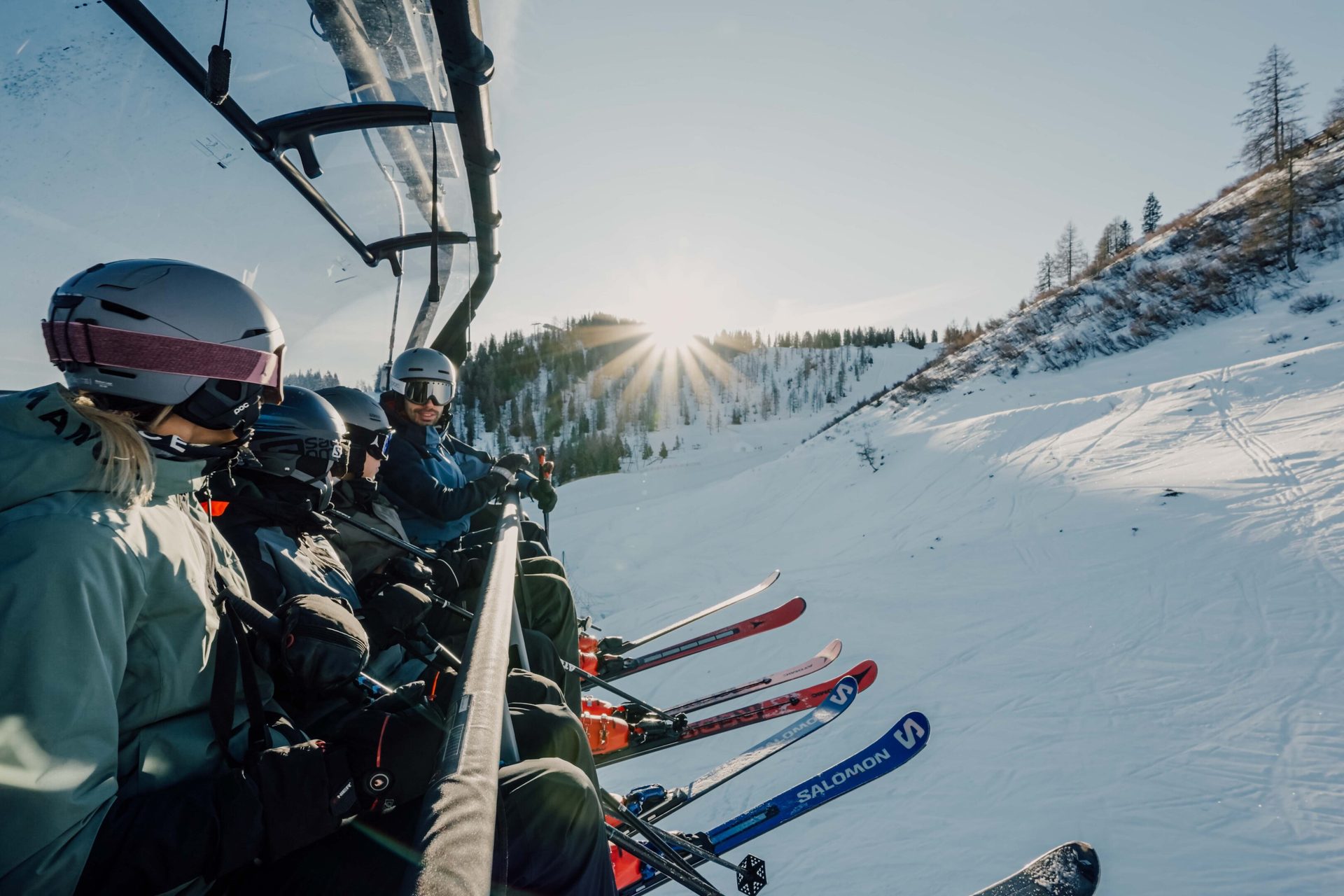Skiers on a chairlift ascend a snowy mountain, with the sun brightly shining over the distant peaks.