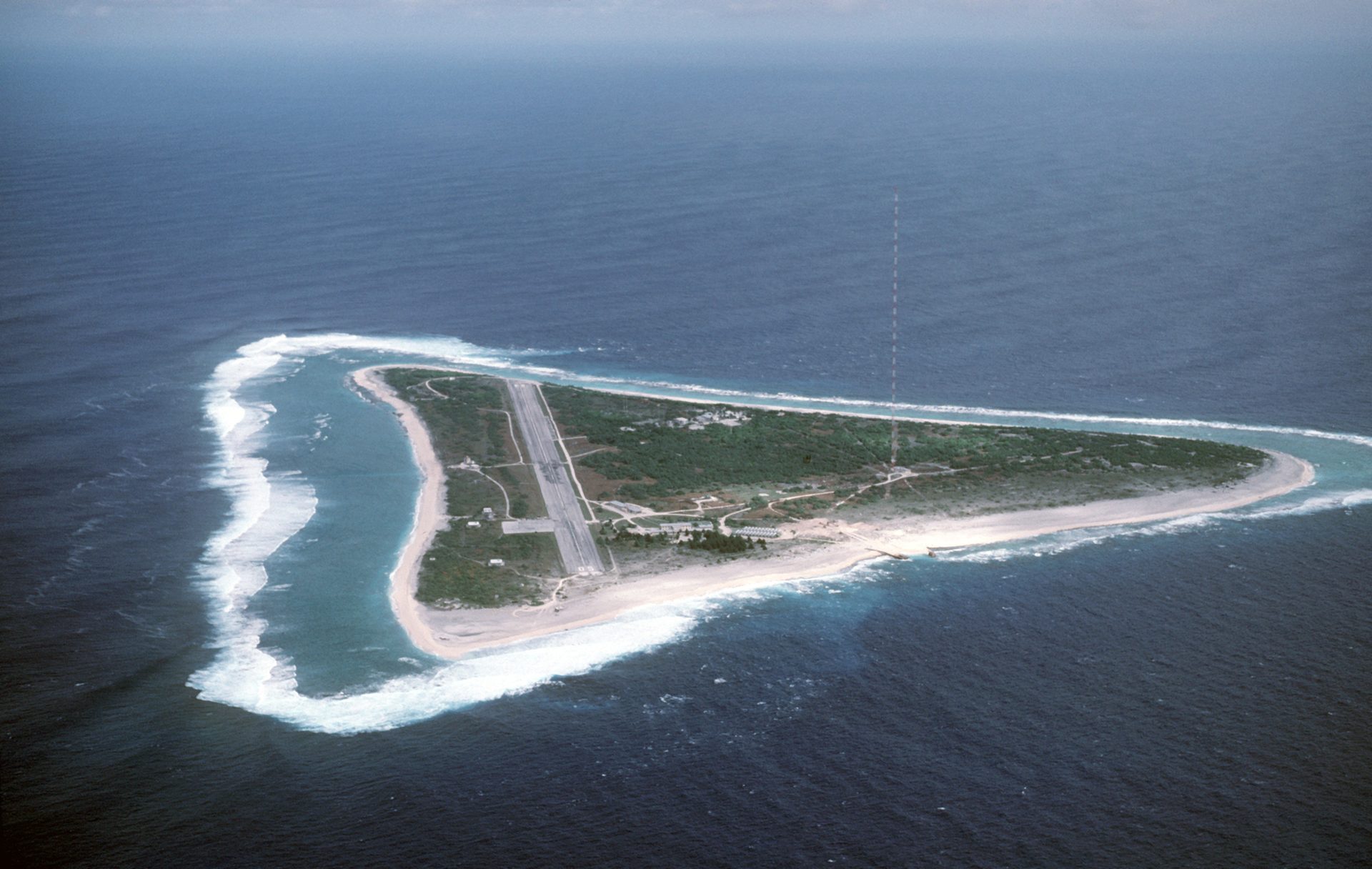 Aerial view of a triangular island with an airstrip, buildings, and a tall tower, surrounded by ocean.