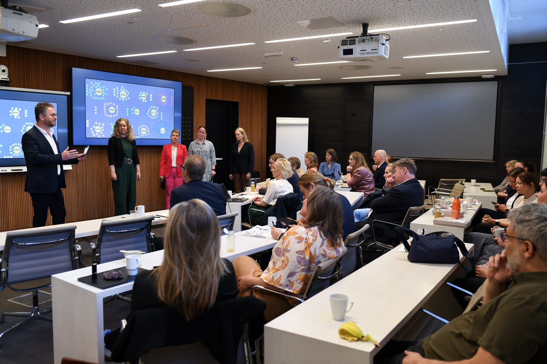 A man presents to an audience in a conference room, flanked by women, with a large screen behind them.