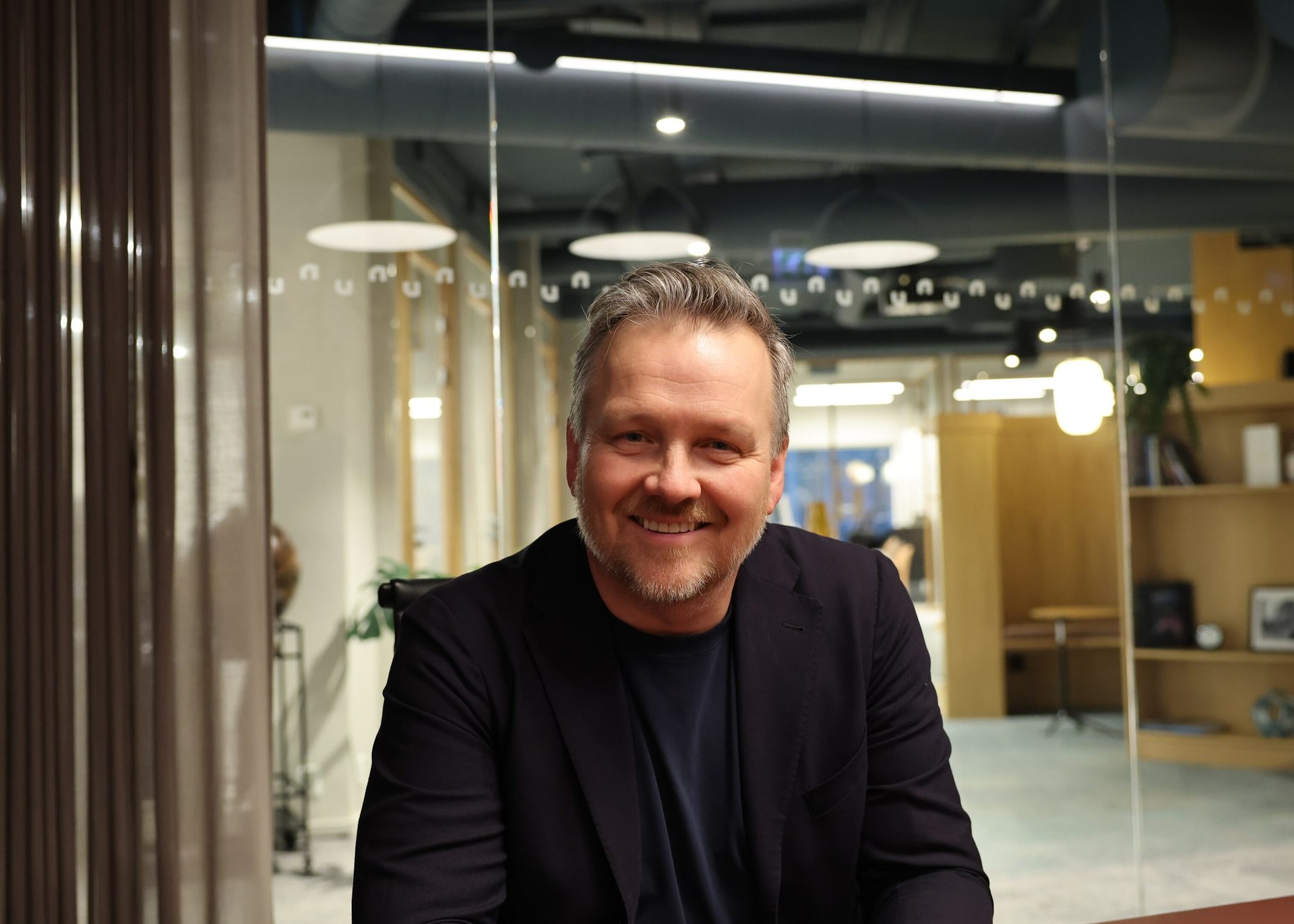A smiling man with graying hair and a beard, wearing a dark jacket over a blue shirt, in an office.