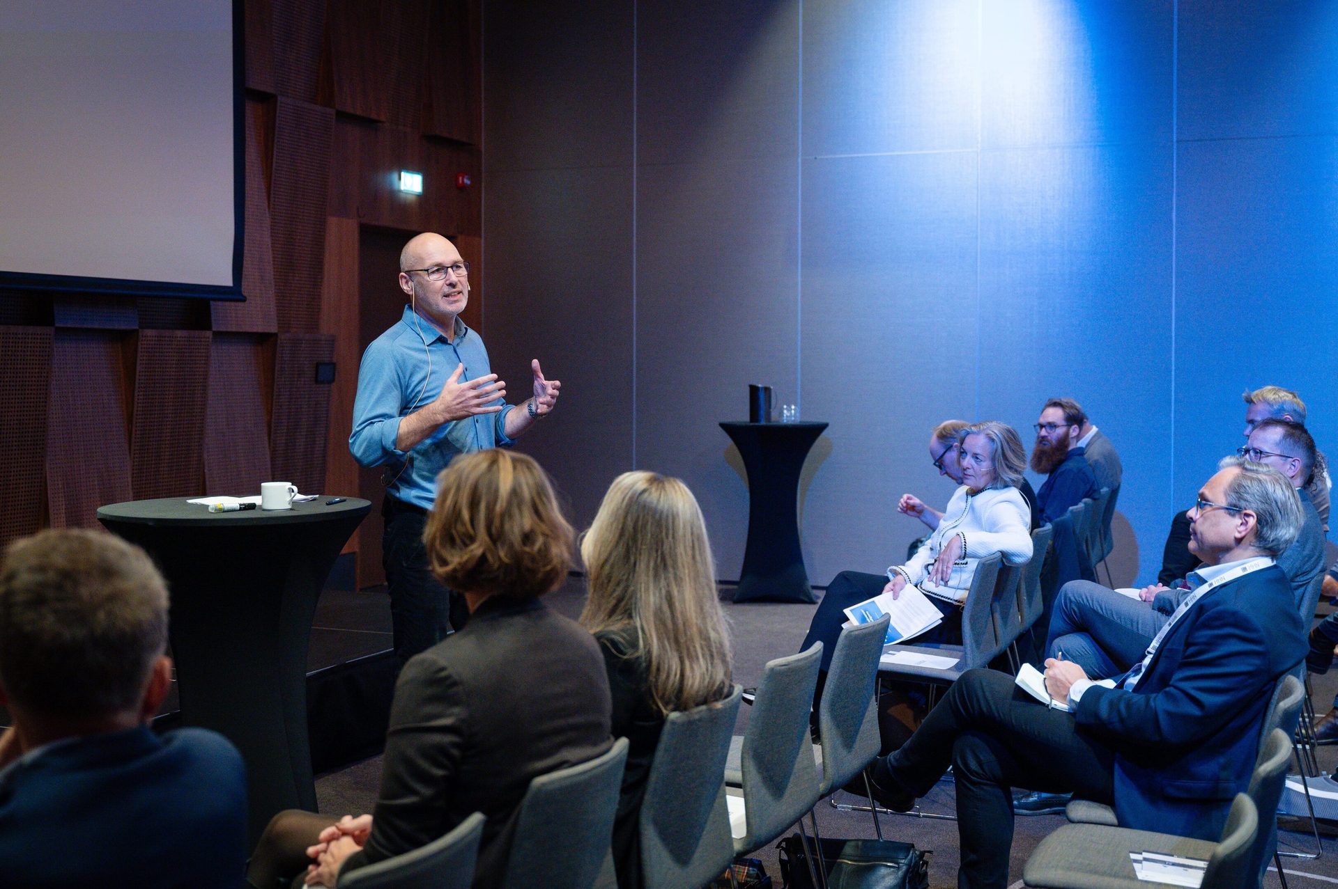 A bald man in a blue shirt presents to an engaged audience in a modern conference room.