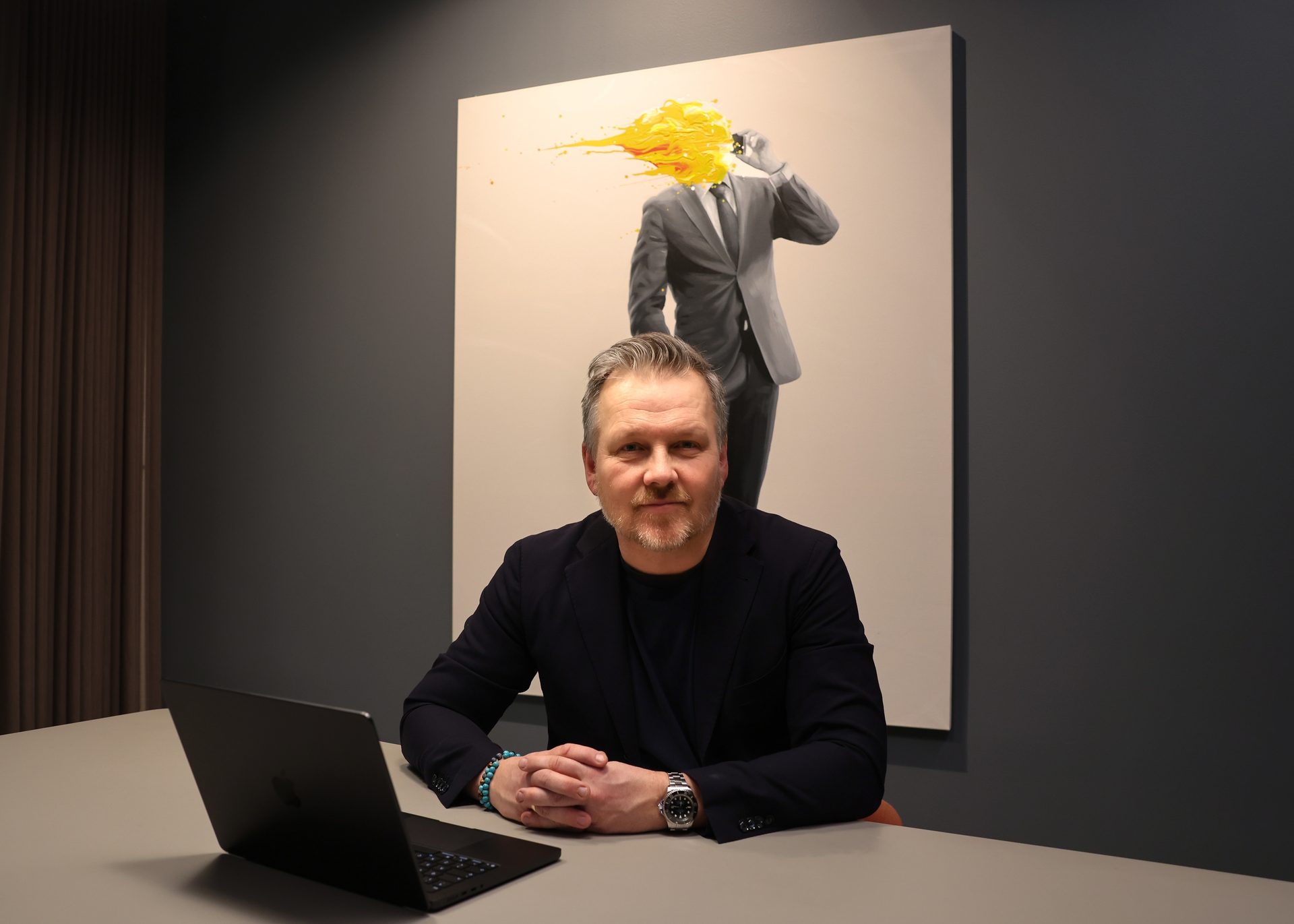 A man with a beard sits at a table with a laptop, in front of a painting of a suited figure with yellow paint.
