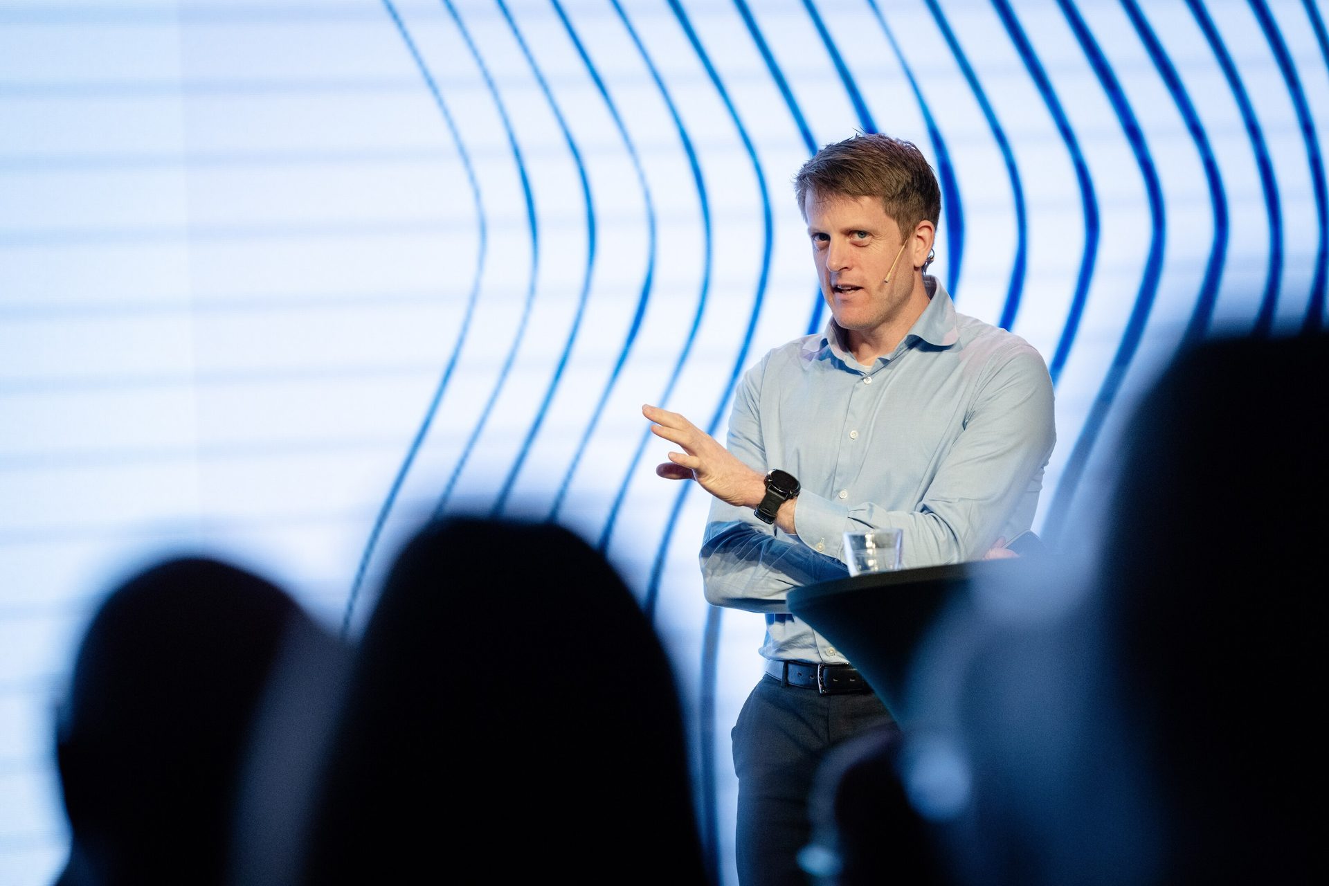 A man in a blue shirt speaking on a stage with a blue wavy background.