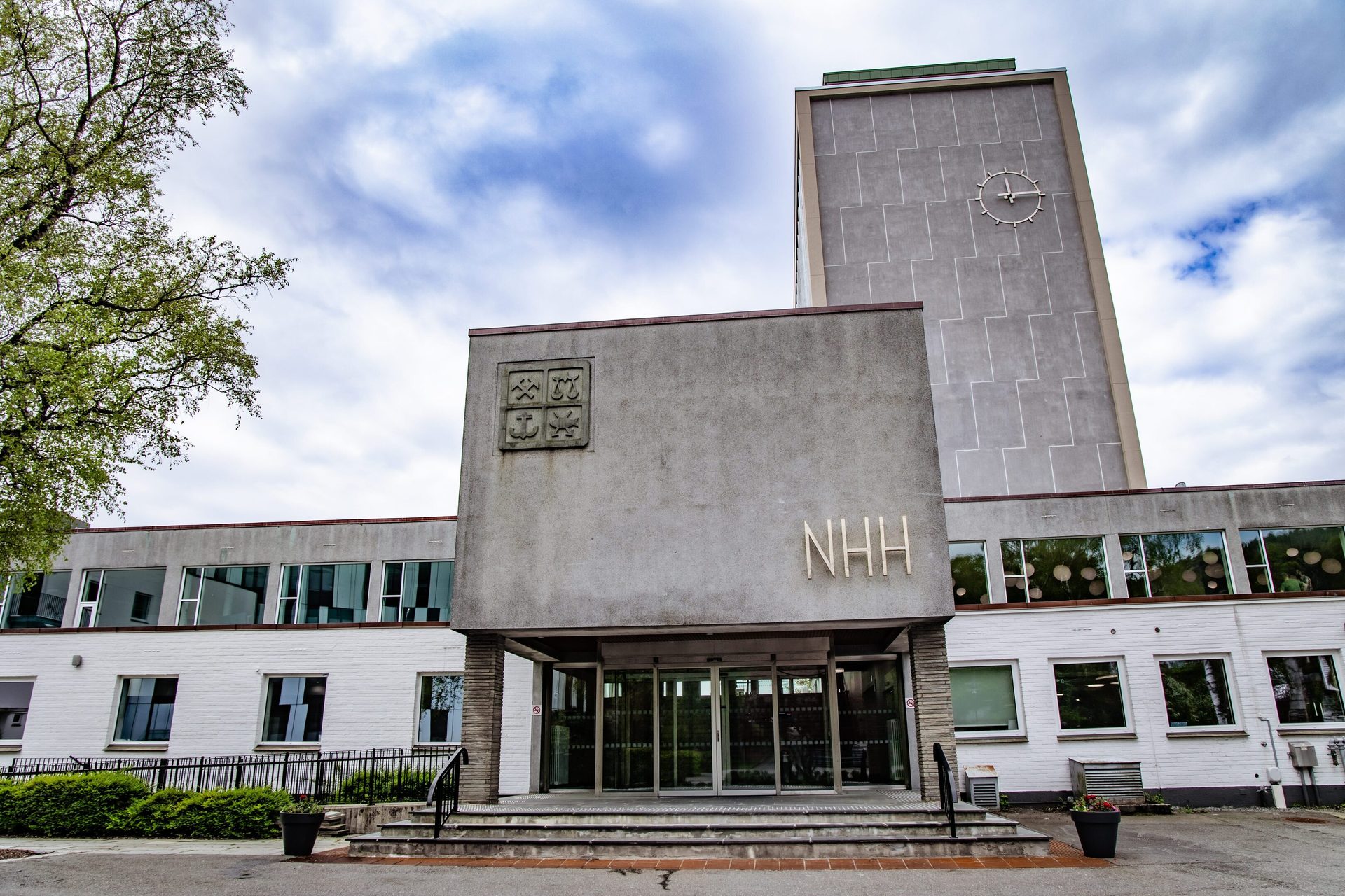 Concrete building with "NHH" sign, a large clock tower with a clock face, and a tree against a cloudy sky.