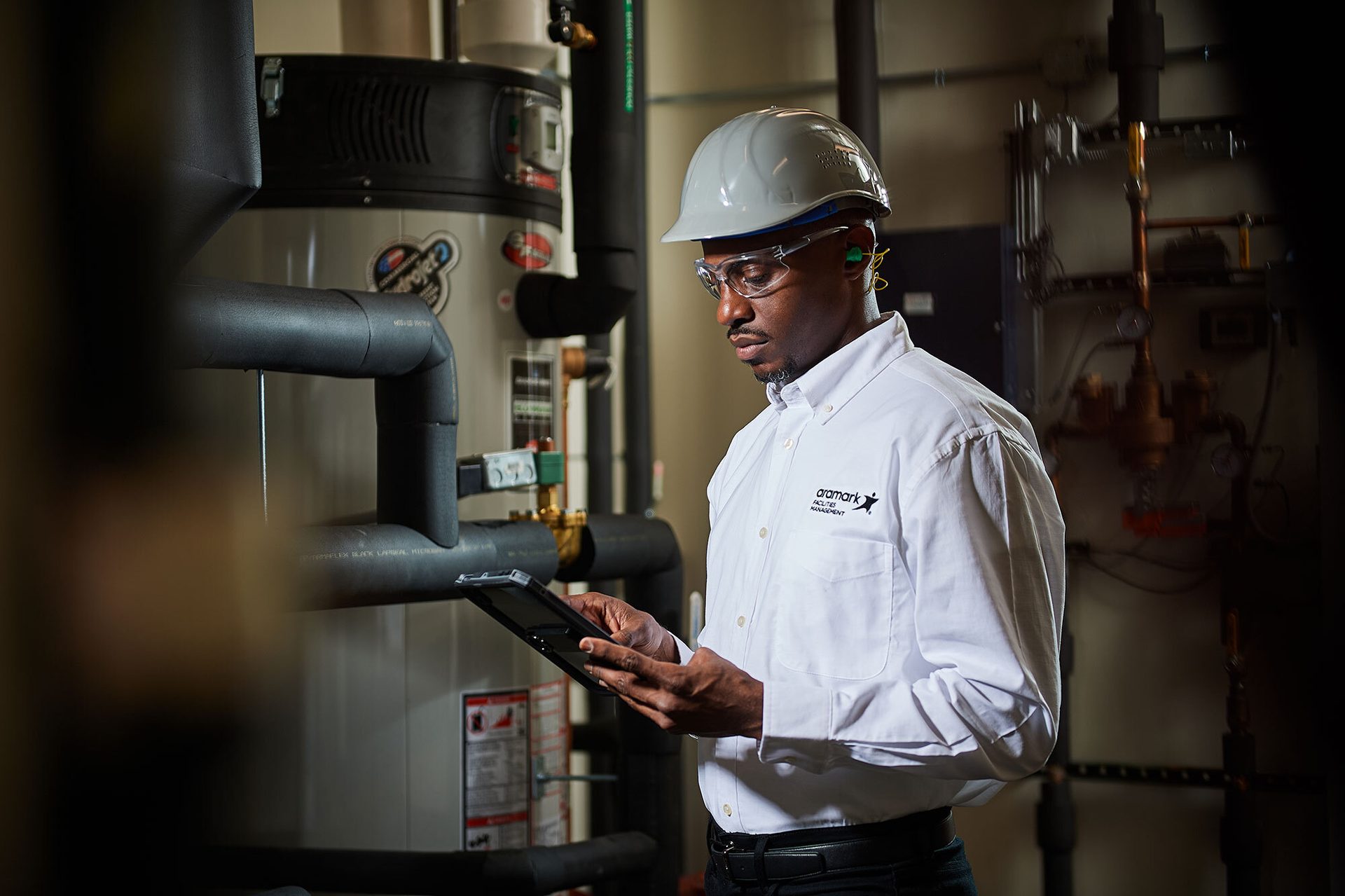 Man in safety gear checking a tablet near industrial equipment.