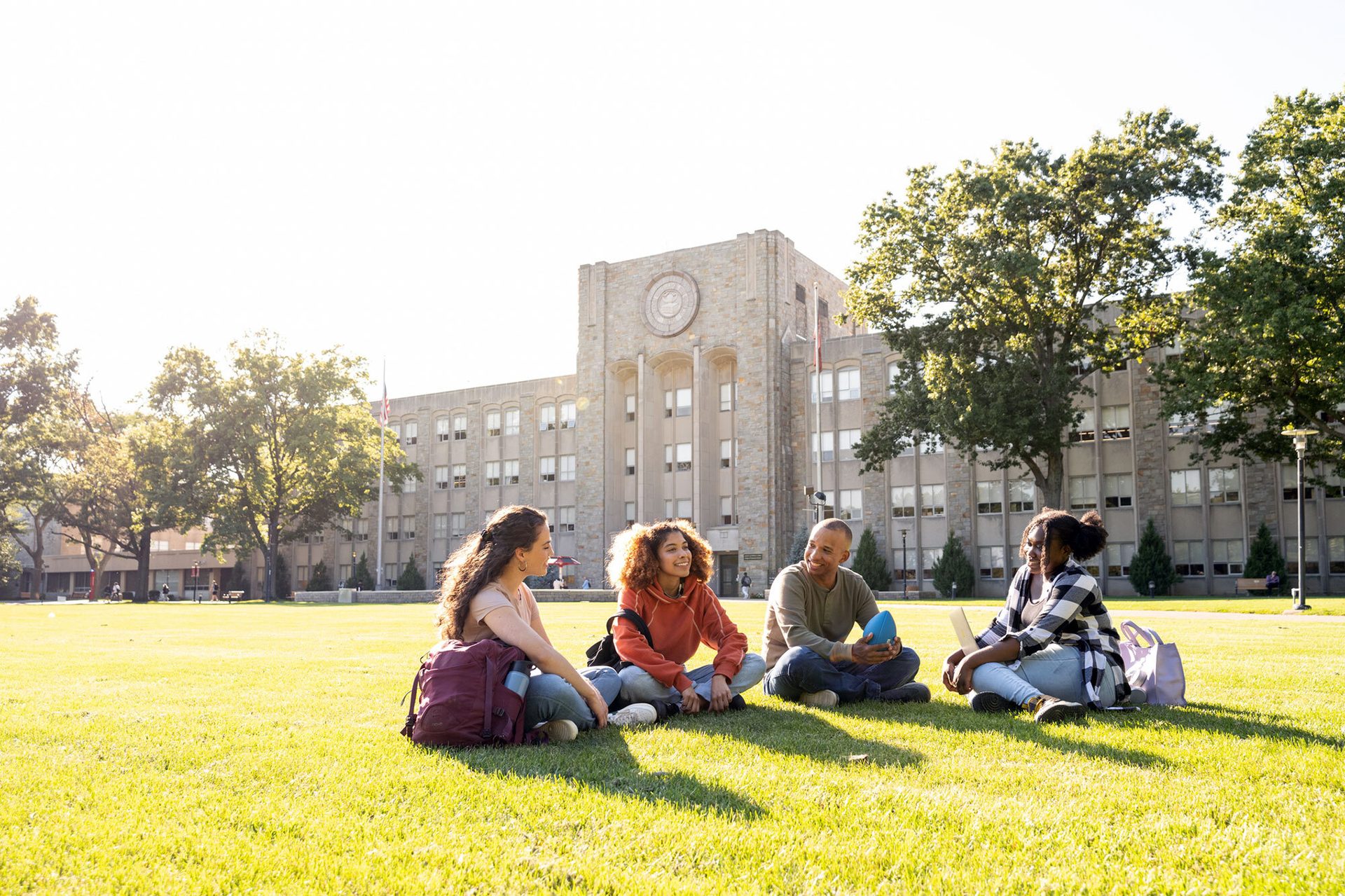 Four students sit on a sunny campus lawn in front of a large building.