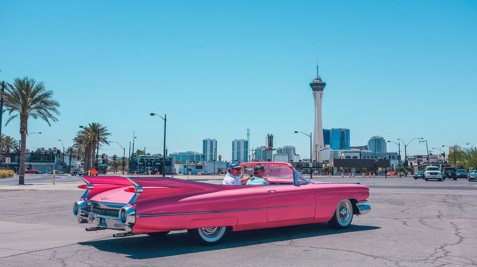 Pink Cadillac convertible with two men, city skyline including Strat tower and palm trees.