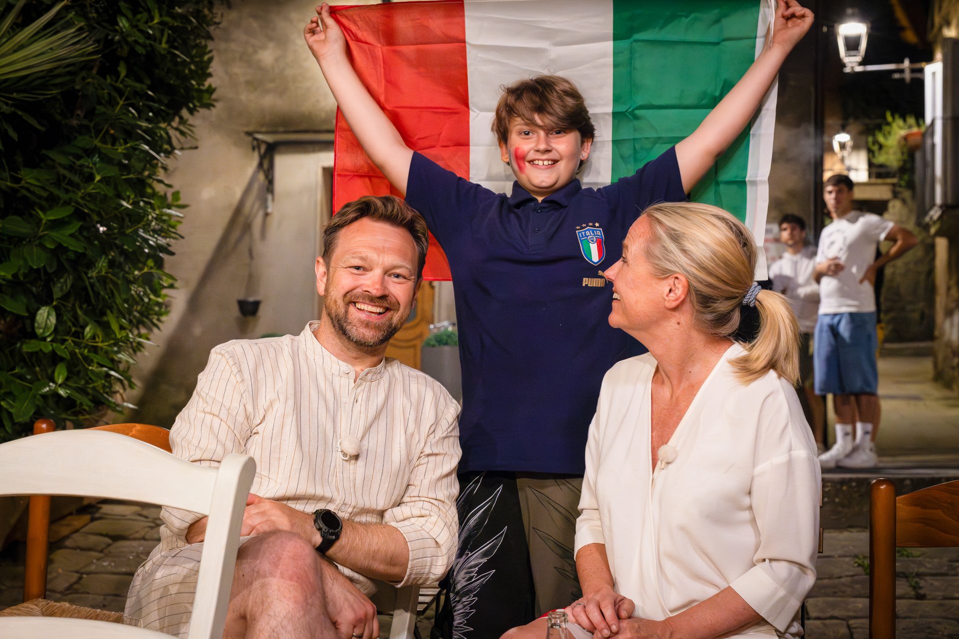 A family, a man, woman, and boy holding an Italian flag, smiles together in a European street at night.