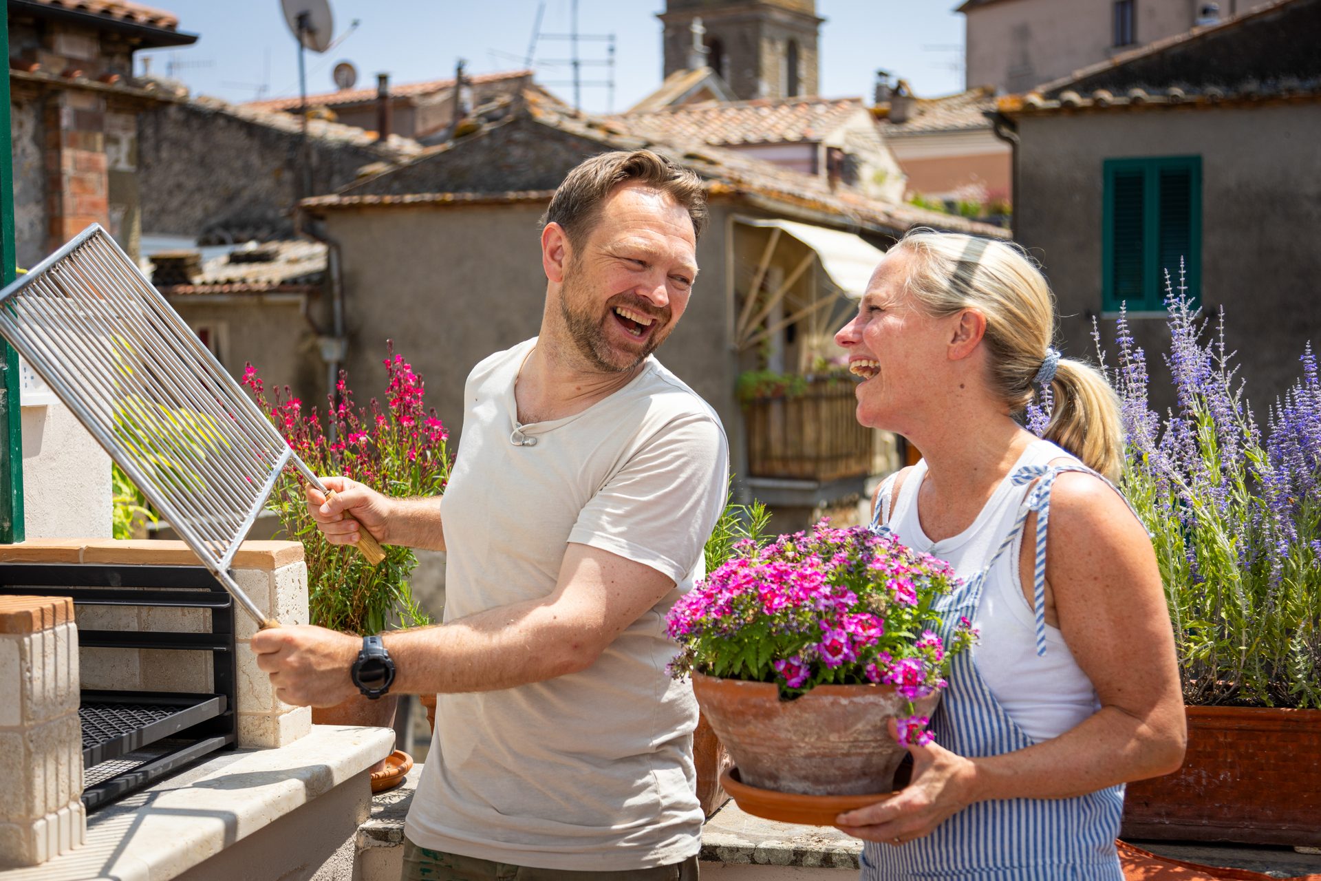 A laughing couple on a rooftop. The man holds a grill grate, the woman holds a pot of pink flowers.