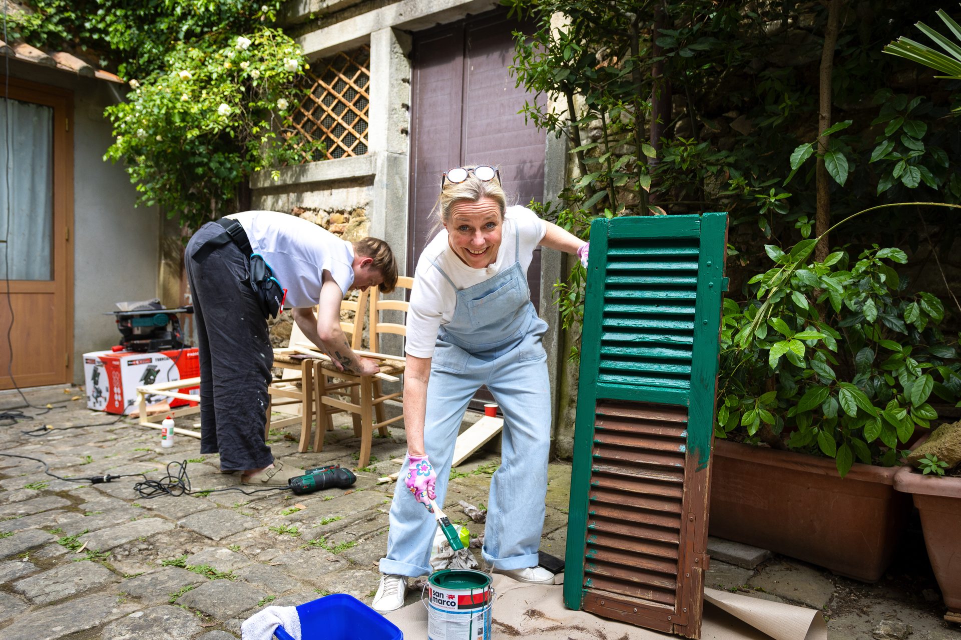 Two people working on DIY projects: a woman painting a shutter green, and a man repairing chairs.