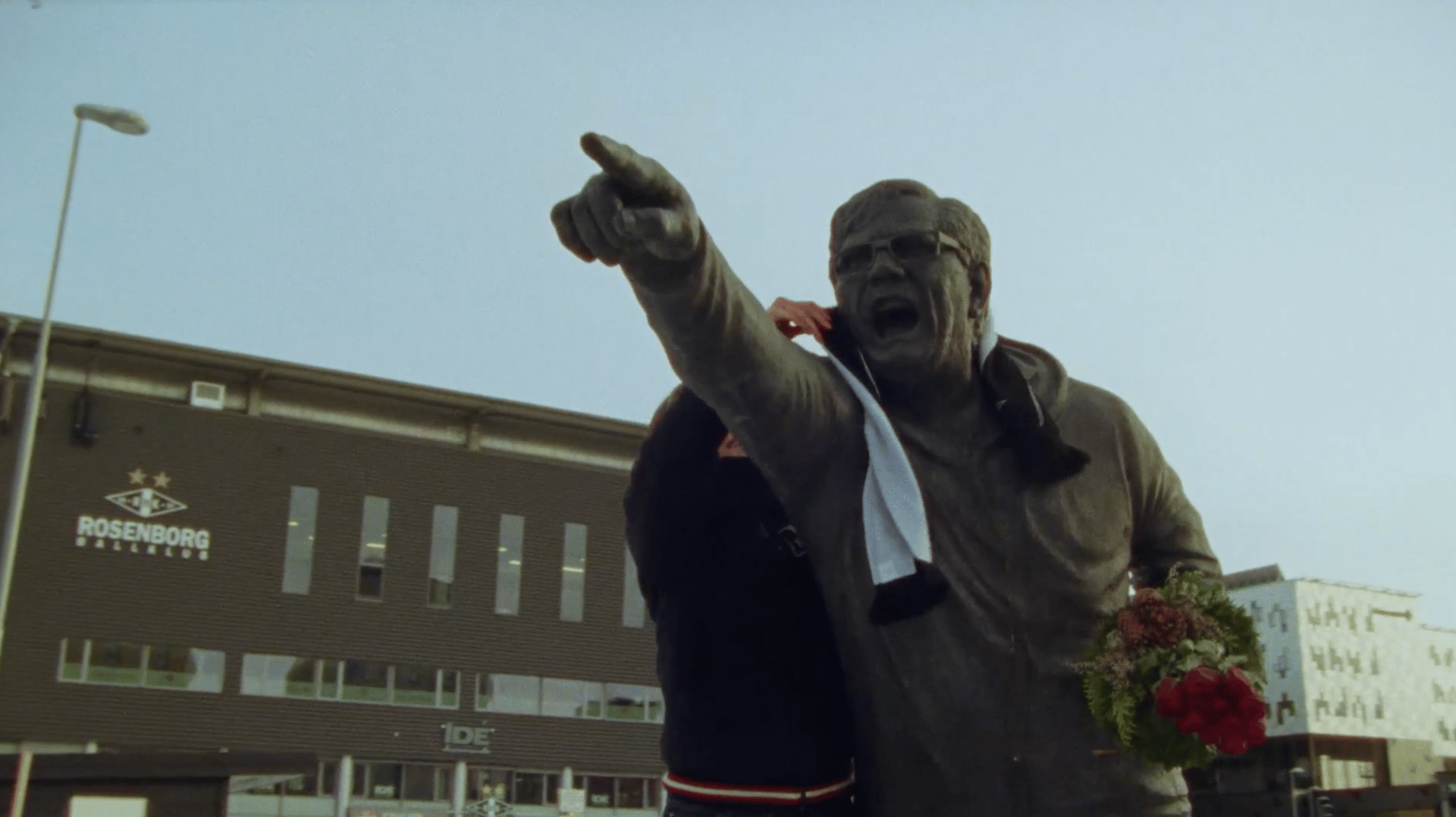 Statue of a man pointing, with a scarf and flowers, in front of the Rosenborg Ballklub building.