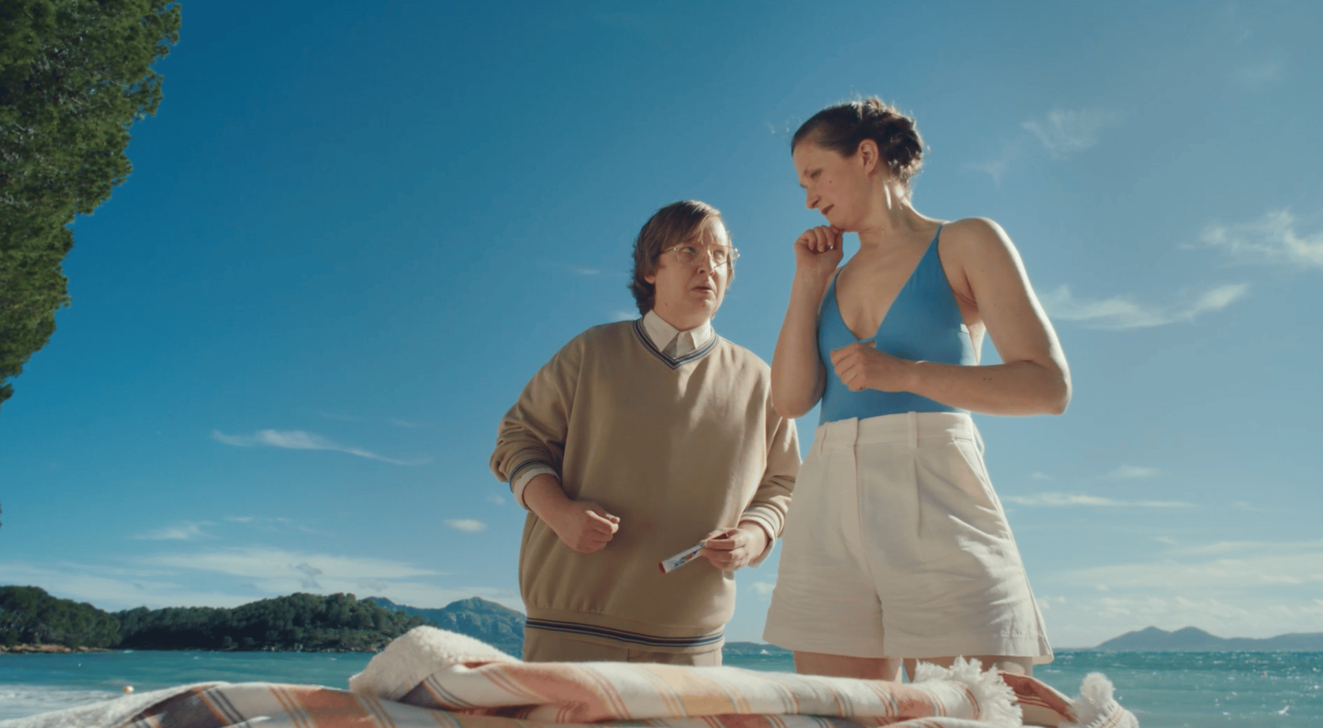 Two people on a sunny beach; a man holds a tube, a woman in a swimsuit looks on.