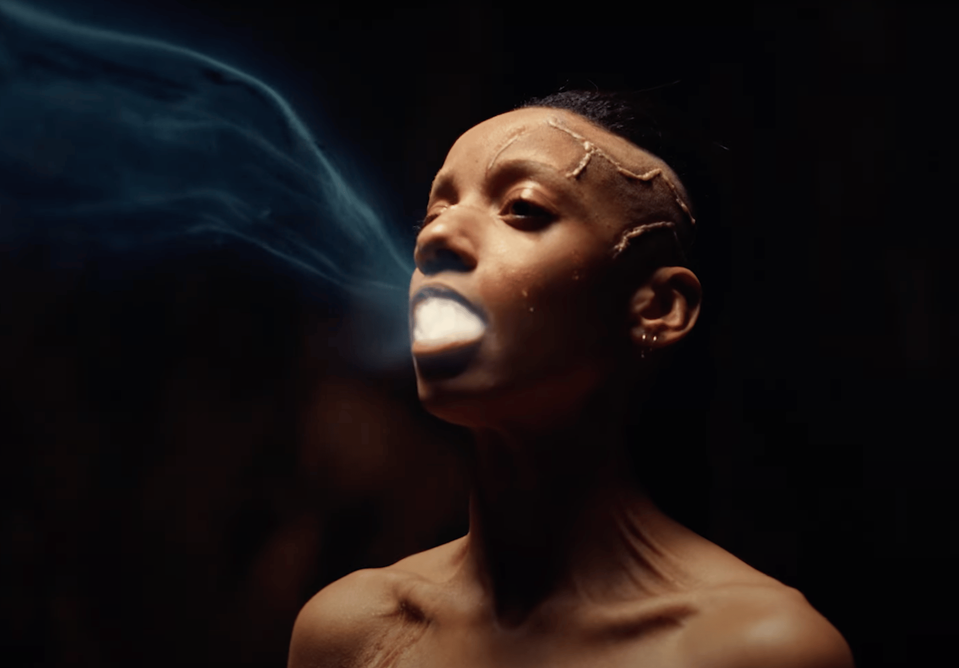 Figure with facial markings exhales luminous blue smoke into darkness.