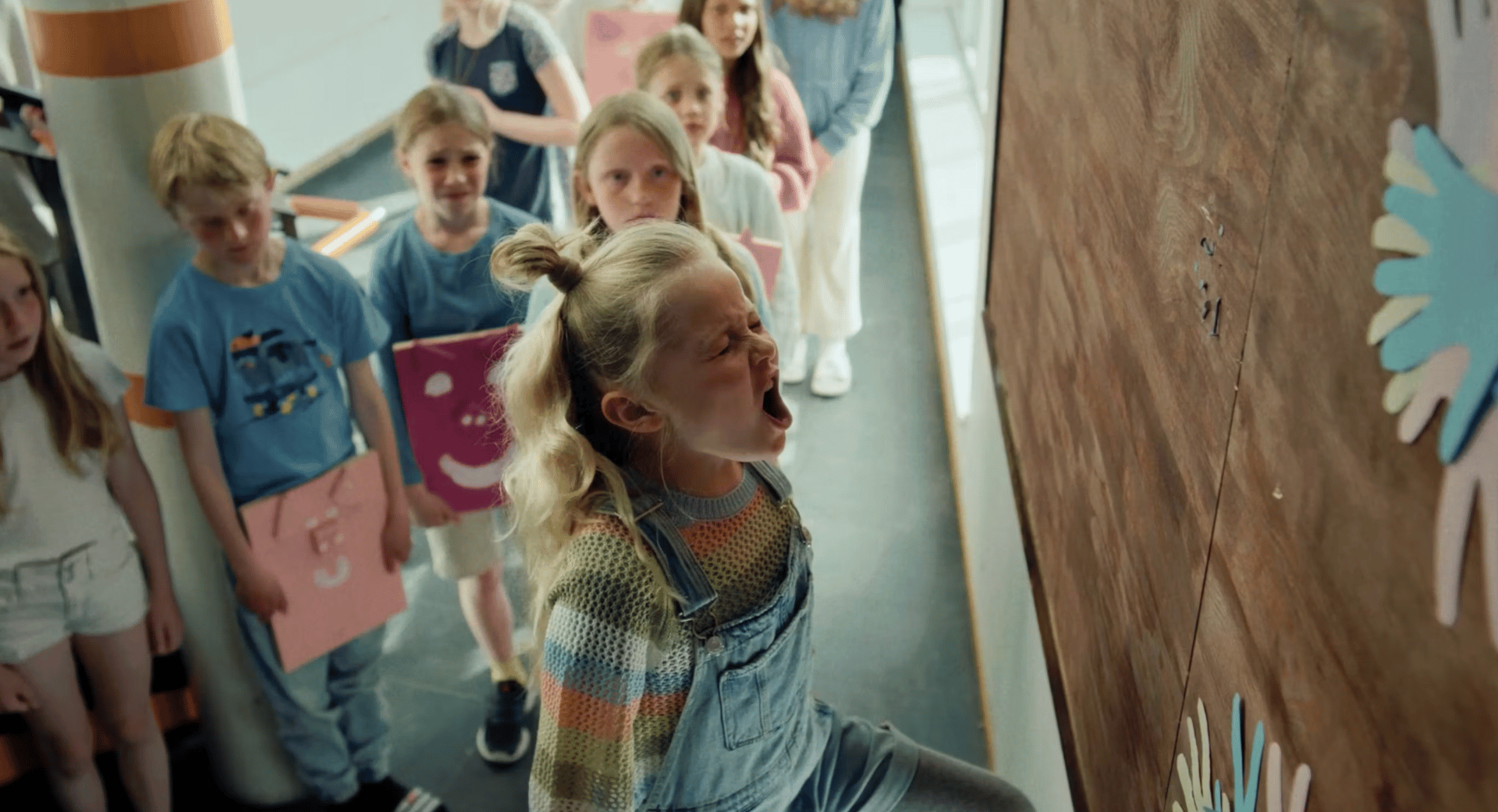 A blonde girl with pigtails loudly screams in a line of children, facing a wooden wall.