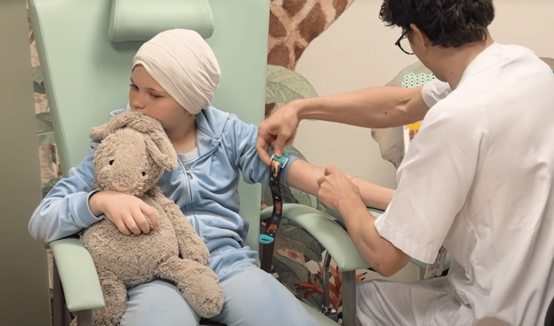 Child in head wrap hugs bunny while medical worker preps arm for procedure.