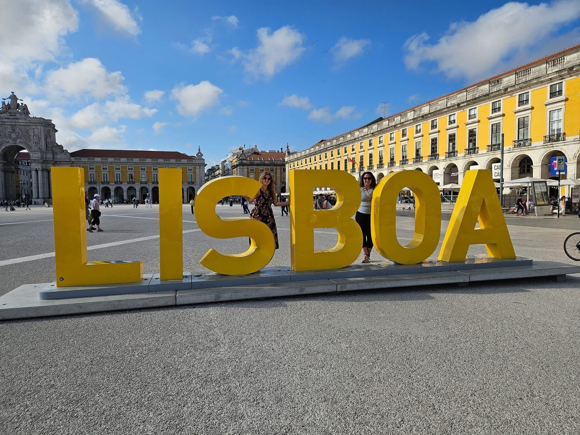 Two women pose with a large yellow "LISBOA" sign in Praça do Comércio, Lisbon.