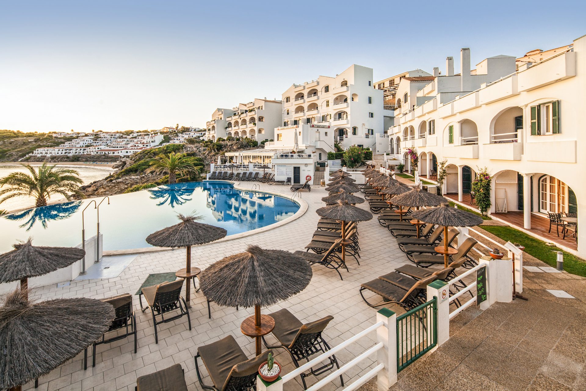 Sunny resort with white buildings, a large pool, and sun loungers overlooking a bay.