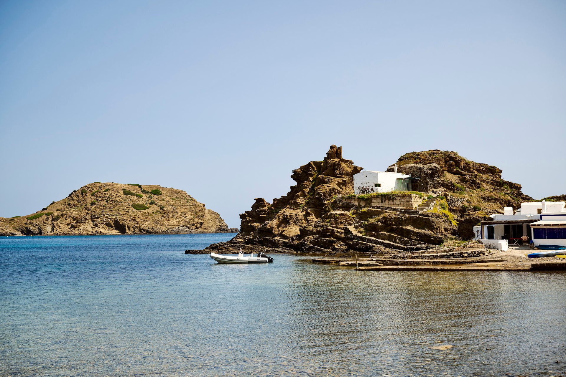 A calm, clear bay with a boat, rocky coast, and white buildings built into the cliffs.