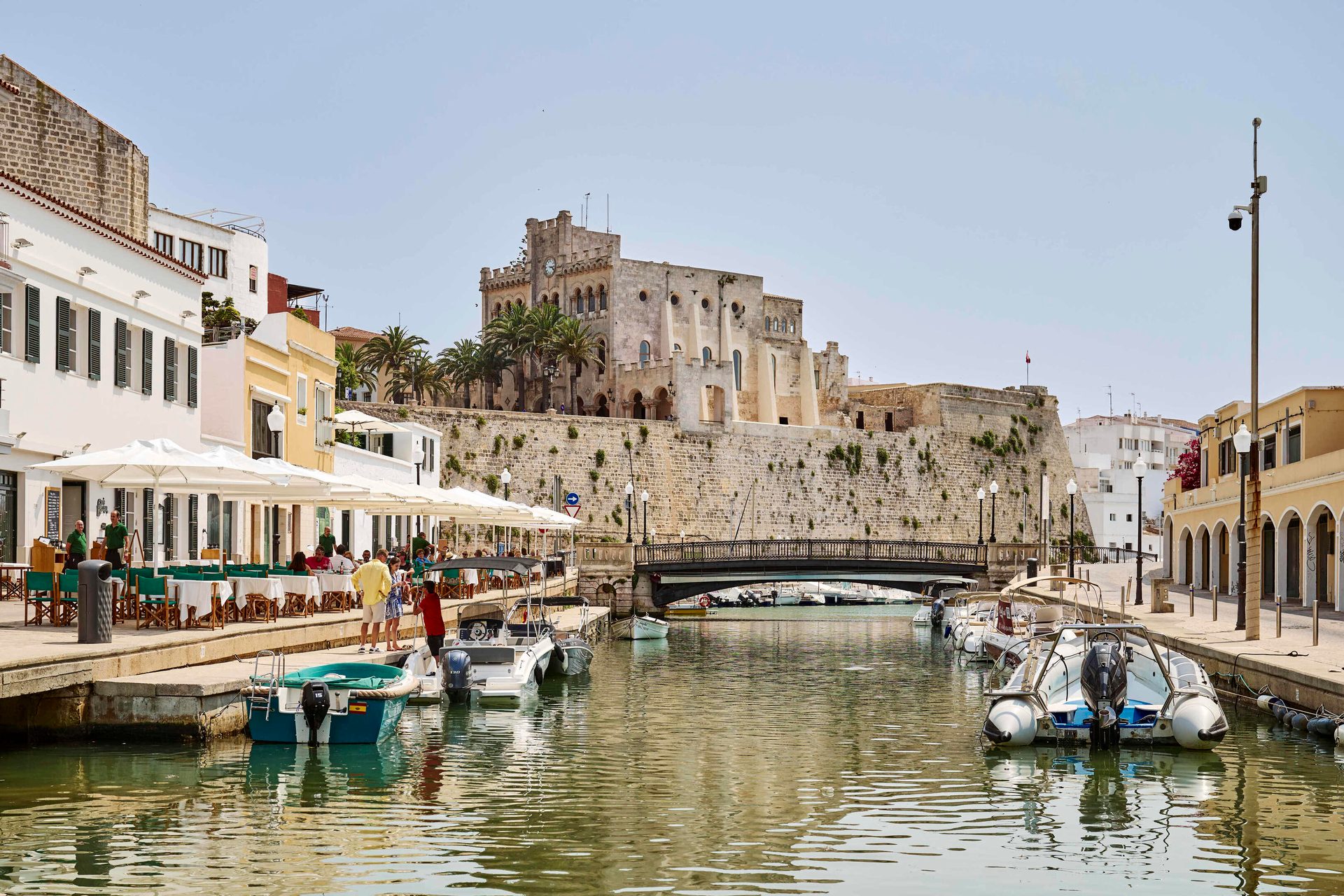 A picturesque canal town with boats, outdoor dining, and a historic building on a stone wall.